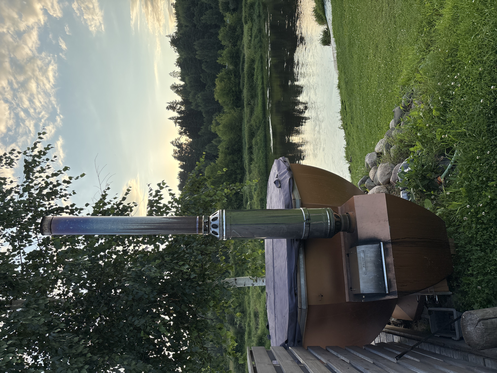 This is a medium shot photograph of a wood-fired hot tub situated outdoors. The hot tub itself is a large, rounded, brown structure with a dark cover. A prominent stainless steel chimney extends vertically from the rear of the hot tub. Below the chimney, attached to the hot tub's exterior, is a metallic firebox with a visible grate. The hot tub is located on a grassy area, adjacent to a wooden fence. Behind the hot tub, a calm river flows, reflecting the sky and the dense green treeline on the opposite bank. The sky is partly cloudy, with soft lighting suggesting either early morning or late afternoon. The overall scene depicts a tranquil, rustic outdoor setting near a body of water. There are no people visible in the image. This scene could be located in a rural area of Zvenigorod, Russia, given the natural landscape and vegetation.
