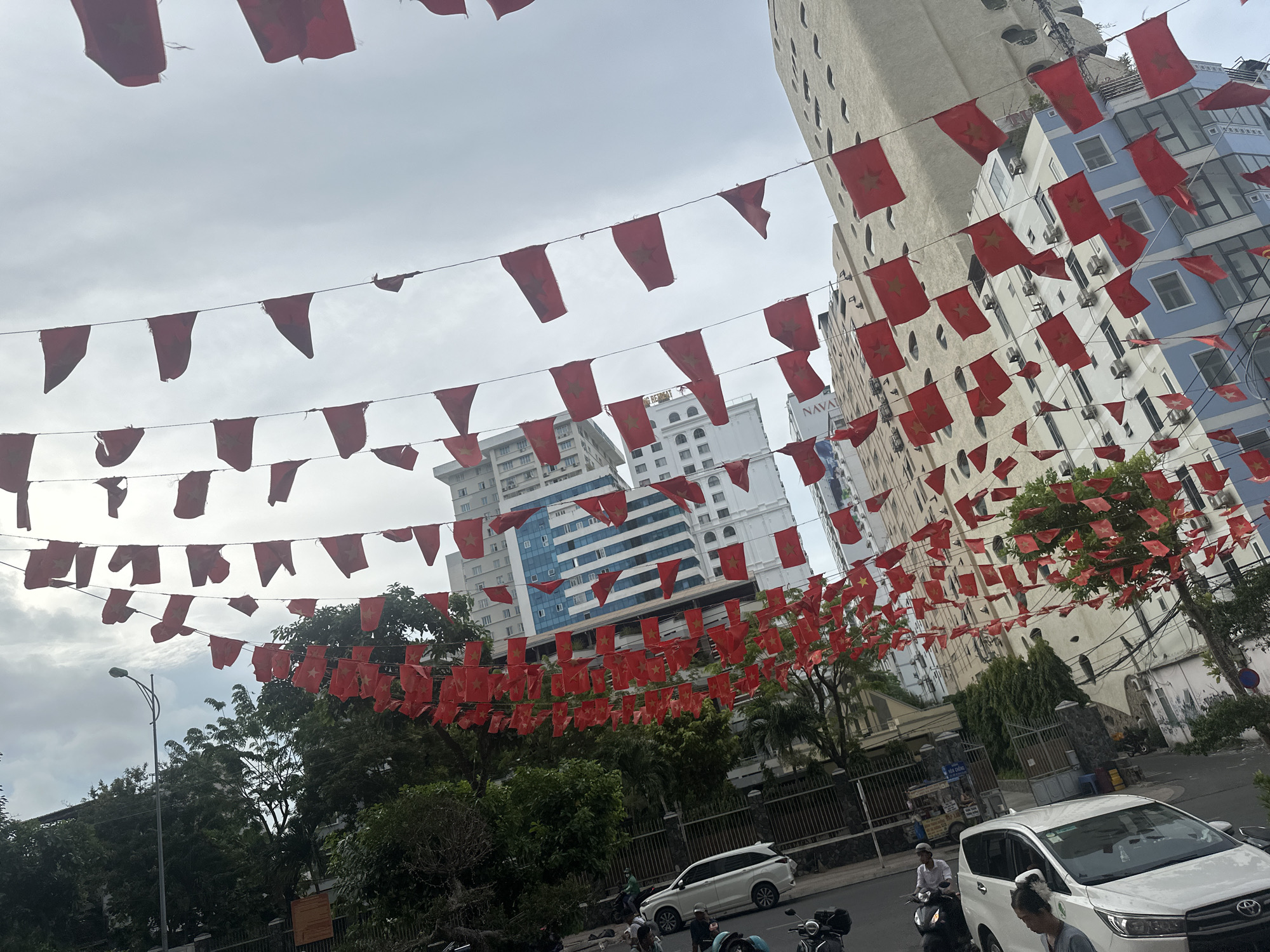 The image shows a street scene in Nha Trang Ward, Vietnam. Red Vietnamese flags hang overhead on strings stretched between buildings. Several buildings are visible in the background, including tall, modern structures with light-colored facades. A street runs through the scene, with several parked cars, motorcycles, and a few people on scooters. The sky is overcast and gray. Trees line the street.