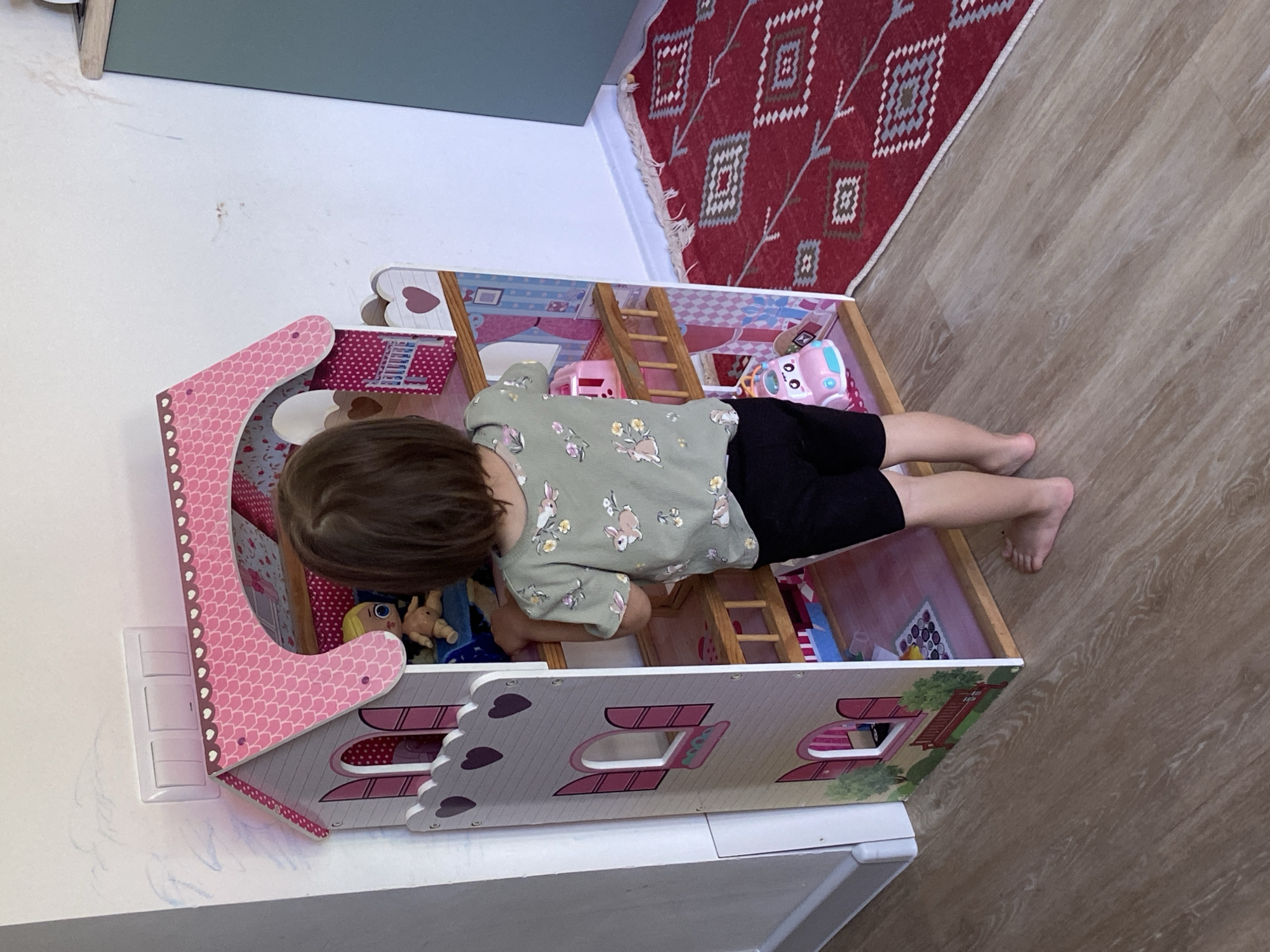 A child is playing with a dollhouse. The child is a young person with short brown hair, wearing a light green t-shirt with a rabbit pattern and black shorts. The dollhouse is pink and white and has multiple rooms. The dollhouse is placed on a white table or shelf. In the background, there is a red rug with geometric patterns and a wooden floor. A wall is visible on the left side of the image, with a light switch. The location cannot be confirmed from the provided images or details.