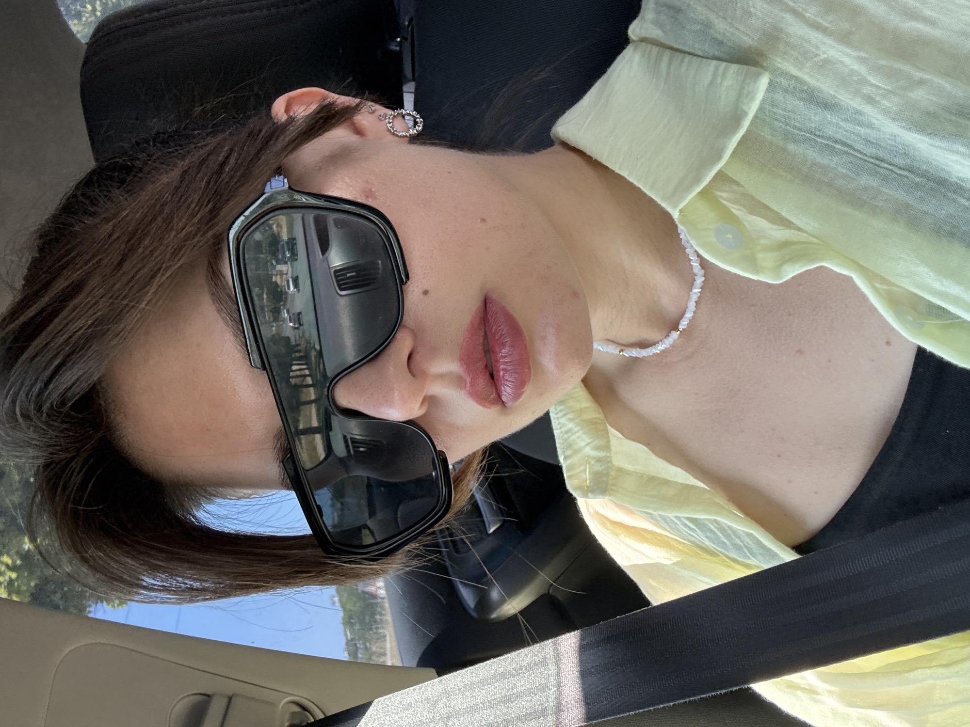 A woman with brown hair pulled back, wearing large, black, wraparound sunglasses and a light yellow collared shirt, is seated in the driver's seat of a car. A black seatbelt is diagonally across her chest. She is also wearing a thin, white beaded necklace and a small, circular earring in her right ear. Her lips are painted with a pink lipstick. The reflection in her sunglasses shows a road with cars and trees. The background inside the car is dark, likely the car's interior, and the window shows a bright, sunny day with trees and buildings outside. The scene appears to be in the city of Unknown, Unknown.