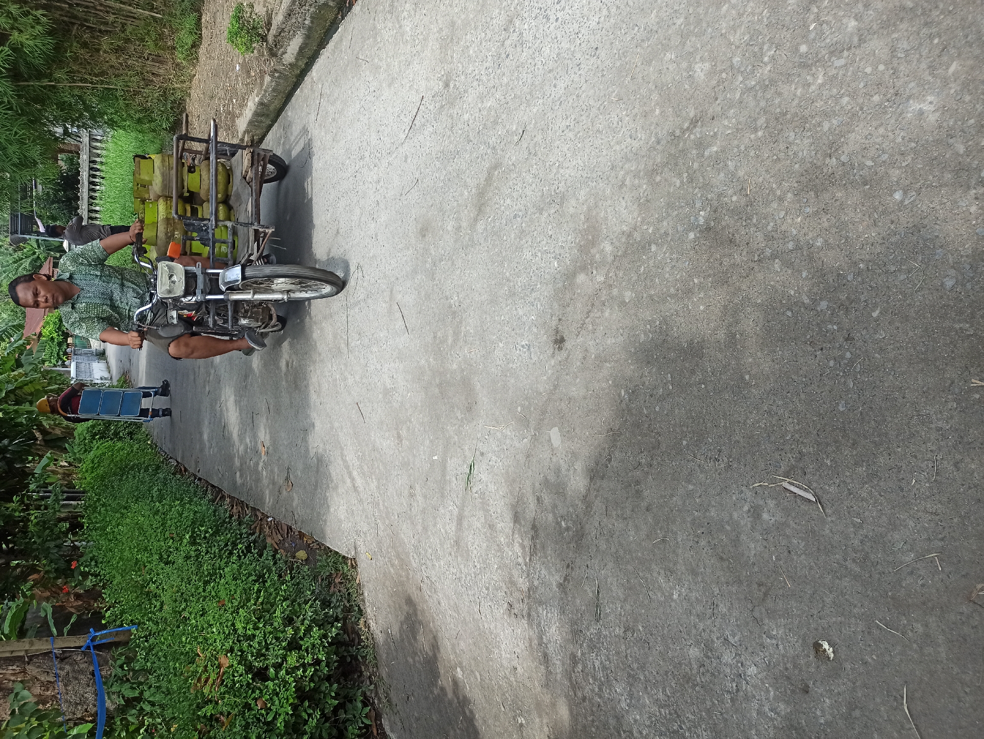 A man is riding a motorcycle with a sidecar in the city of Medan, Indonesia. The motorcycle is carrying several green propane gas cylinders in the sidecar. The man is wearing a short-sleeved patterned shirt and shorts. The motorcycle is on a paved road. To the left of the road is a dense green hedge. To the right of the road is a drainage ditch and some bamboo plants. In the background, another person is visible pushing a cart. The scene appears to be in a residential area.