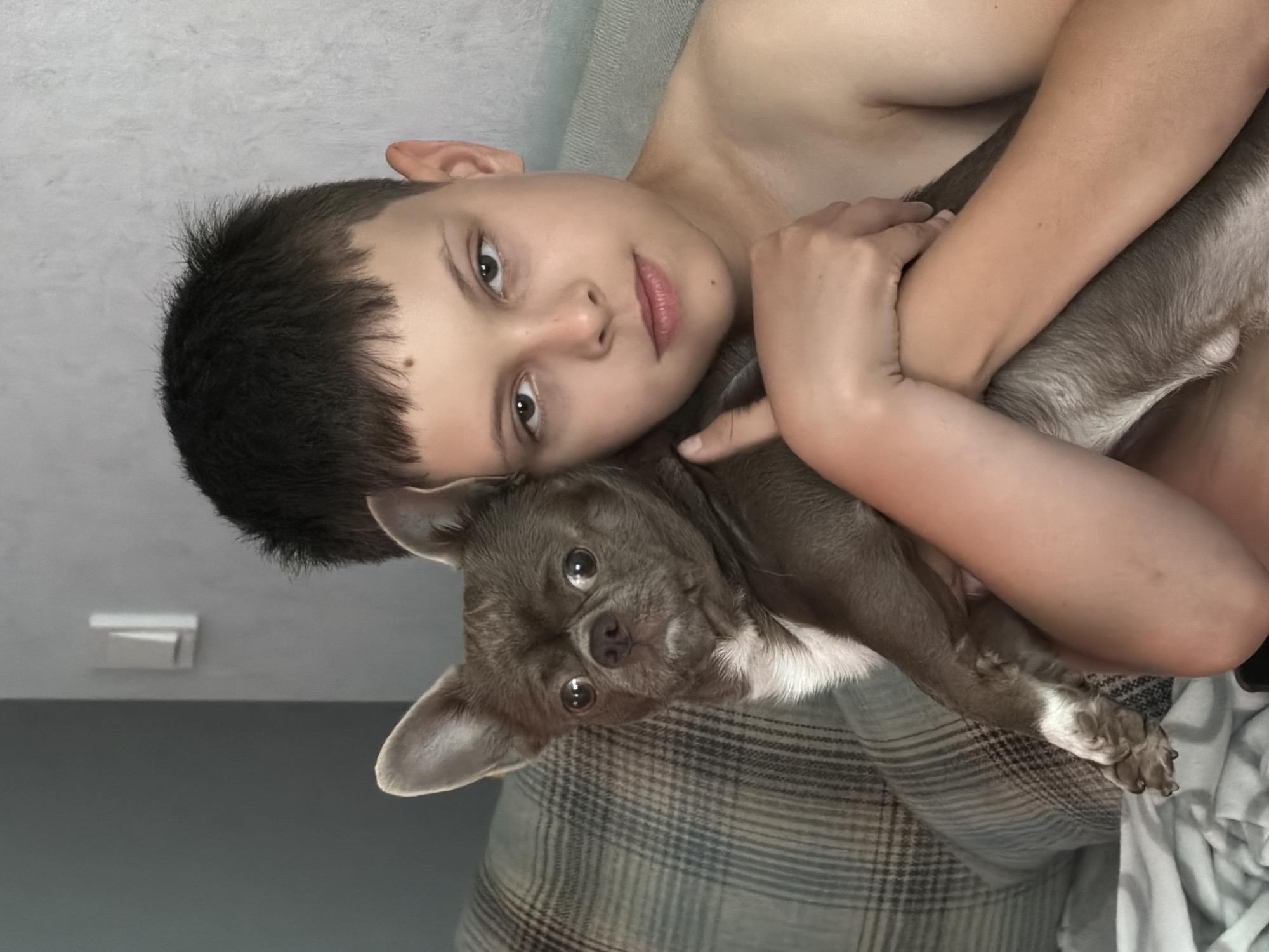 The image features a young person and a small dog, likely a chihuahua, in a close embrace. The person, a boy with short dark hair and light skin, is lying down and holding the dog. His head is tilted, resting on the dog or a surface, and he is looking towards the camera with a gentle expression. The dog, with large expressive eyes and light brown fur, is nestled in the boy's arms. The scene appears to be indoors, as suggested by the soft lighting and the background elements, which include a textured wall or surface and a plaid blanket. The overall mood is one of affection and comfort between the boy and his pet. The image is oriented sideways. No text is visible in the image. The location context is Votkinsk, Russia.