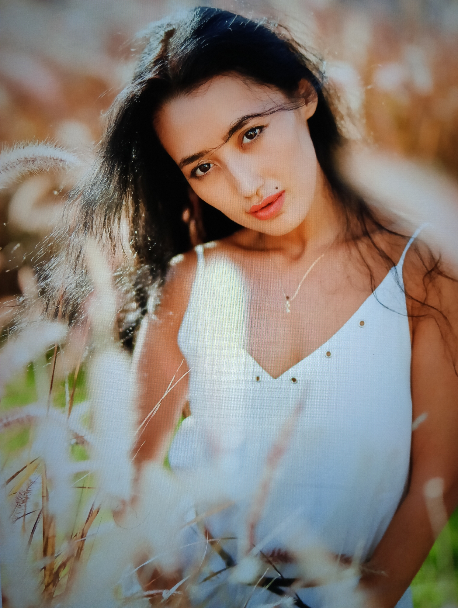 A young woman poses outdoors in a field of tall grass. She has long, dark hair and is wearing a white, sleeveless top with small metallic details. She wears a delicate necklace. The background is blurred, suggesting a shallow depth of field. The scene is bathed in soft, natural light. The image may be taken in a scenic area near Nha Trang Ward, Vietnam.