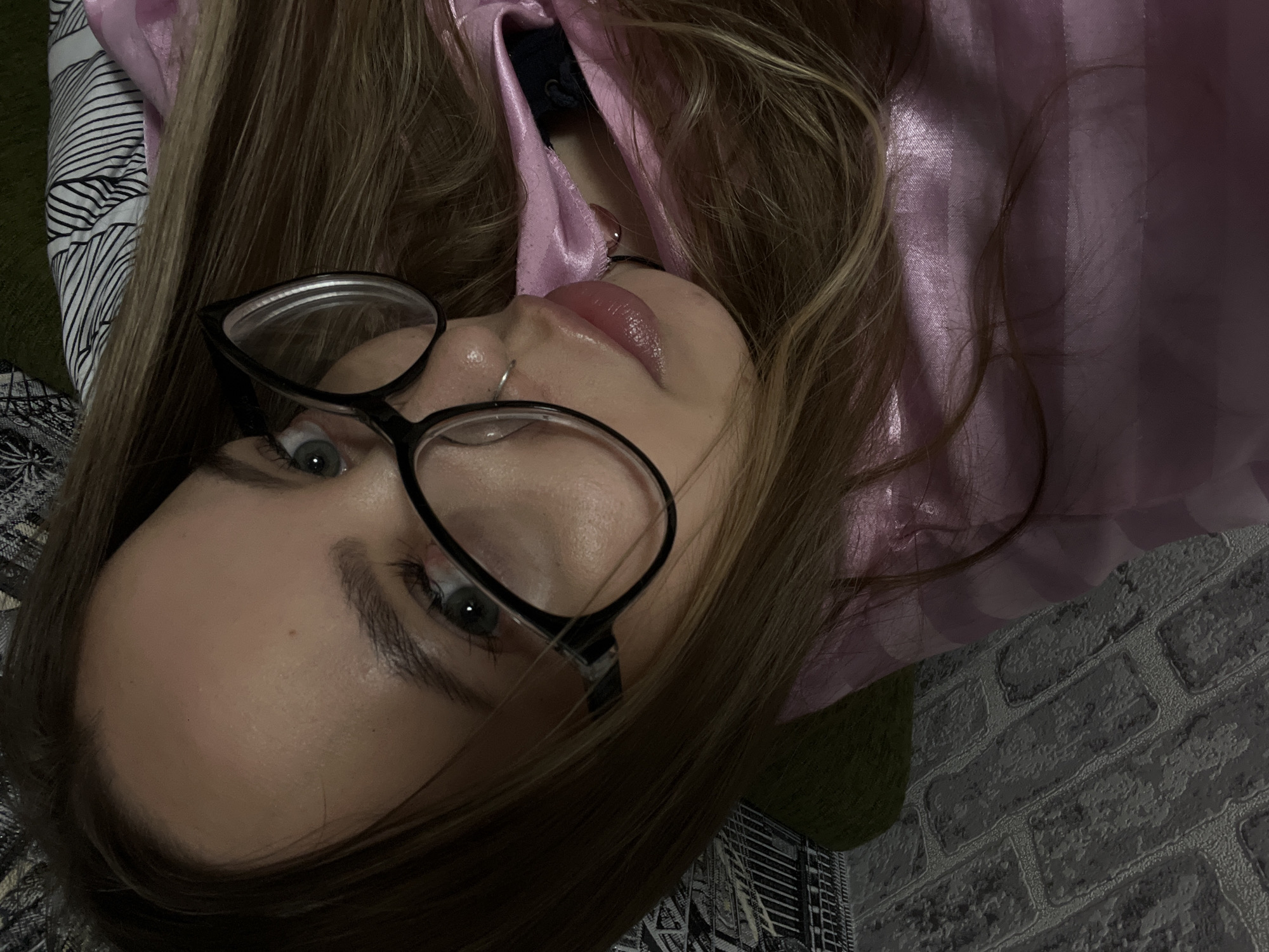 A woman with fair skin and light brown hair is lying on her back. She is wearing black-framed glasses and a pink satin top. She has a nose ring. The background is a mix of textures including a patterned fabric, a green surface, and a brick-patterned wall. The image does not contain enough information to determine a specific location in Novopokrovskaya, Russia.