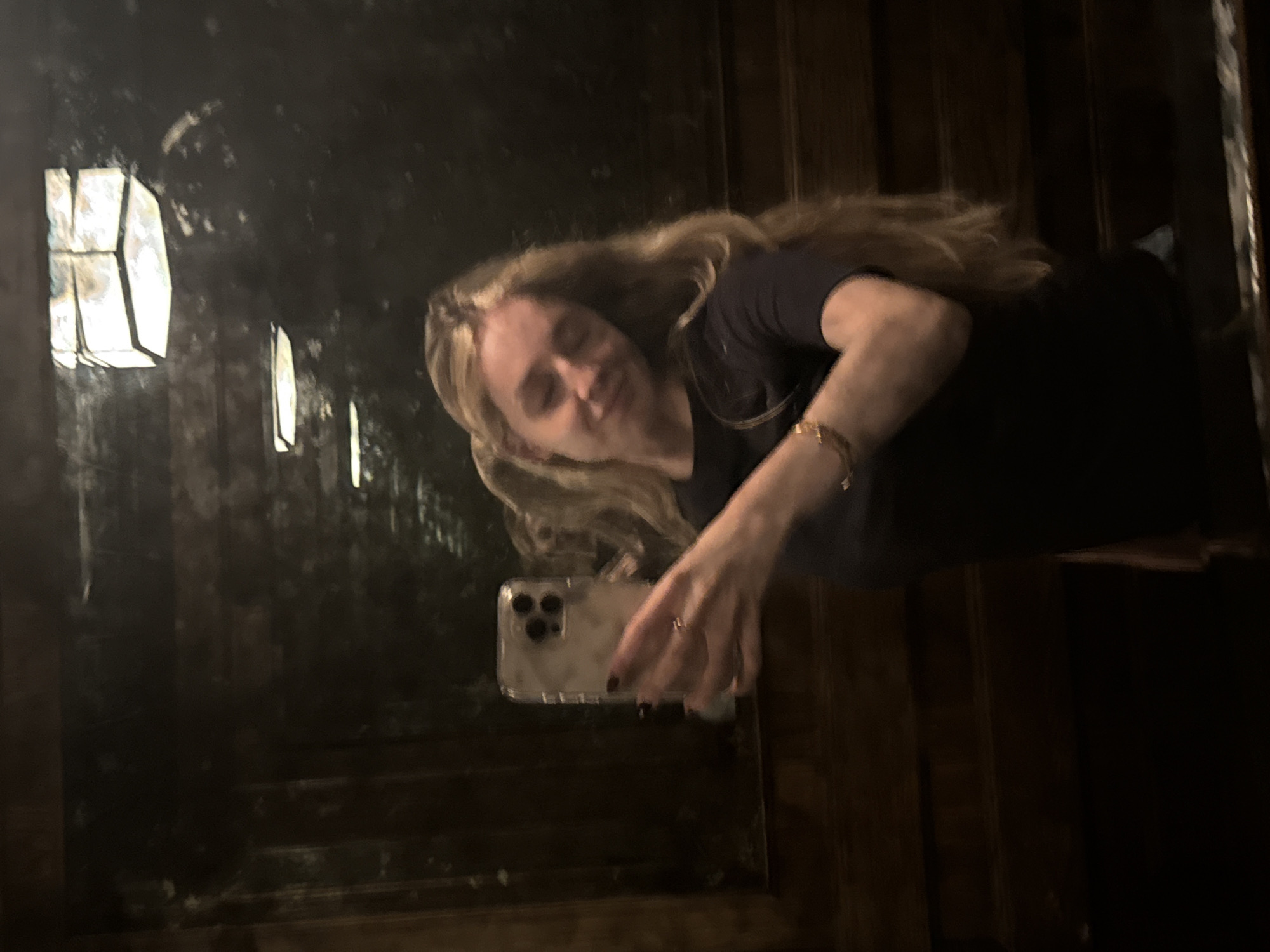 A woman with blonde hair is taking a selfie in a mirror. The reflection shows a row of windows. The environment is dark, and the location could be in Mexico City, Mexico.