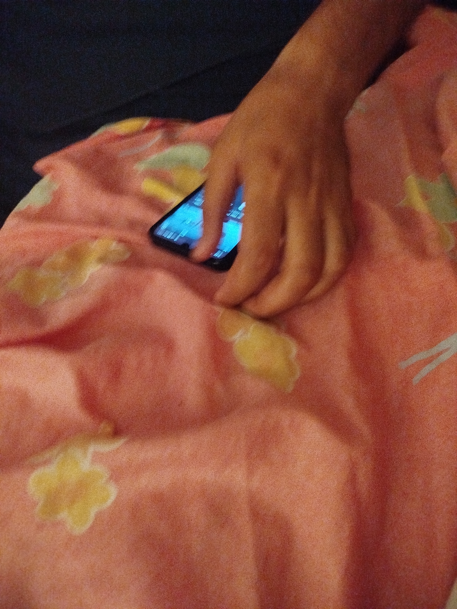 A person's hand is shown resting on a pink blanket with a floral pattern. A smartphone is visible beneath the hand, its screen illuminated with blue light. The background is dark and appears to be a surface like a couch or bed. There is no information in the image to suggest a location in Lassan, Pakistan.