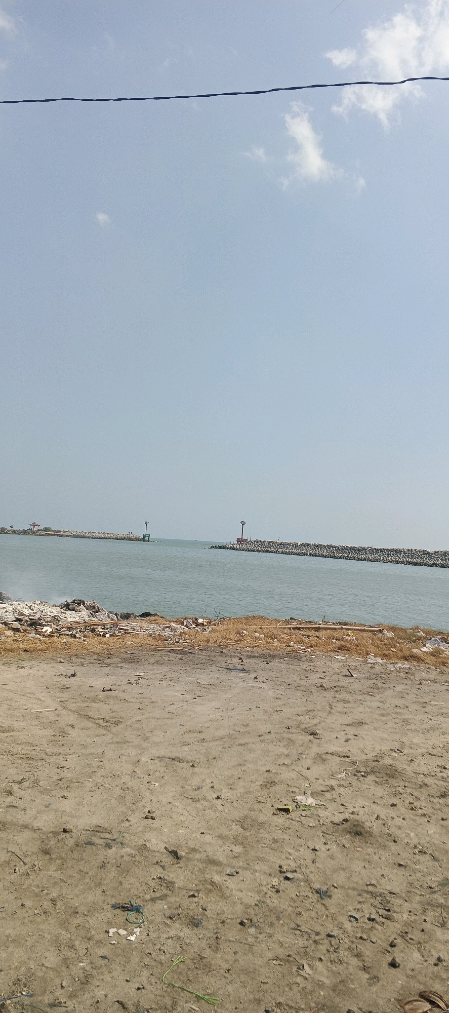 The image captures a view of a coastal area, likely a harbor entrance, with a breakwater made of large rocks extending into the sea. Two lighthouses, one red and one green, mark the entrance. The water is calm and a pale blue, reflecting the clear sky above. In the foreground, there is a patch of dry, sandy ground littered with debris, including pieces of wood, plastic, and other refuse, suggesting a neglected or undeveloped shoreline. A single dark wire stretches horizontally across the top of the frame, obscuring a portion of the sky. The scene is devoid of people or any obvious human activity, presenting a quiet, somewhat desolate, natural and man-made environment.