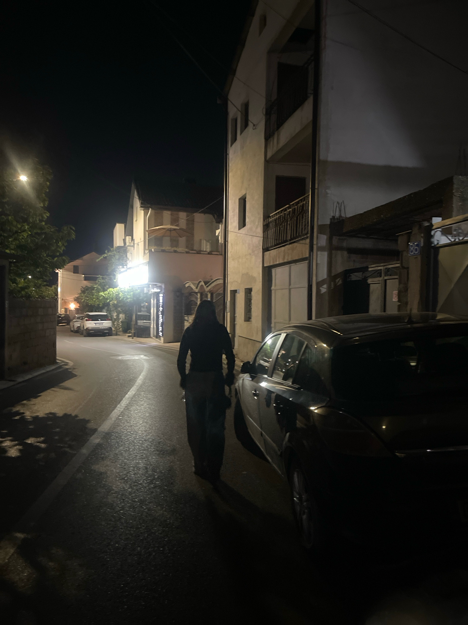 A person walks down a dimly lit street at night in Prizren, Kosovo. The street is paved with asphalt and has a white line marking the center. Buildings line both sides of the street. A dark-colored car is parked on the right side of the street. Another white car is parked further down on the left side. Trees and streetlights are visible. The person is wearing dark clothing and appears to be walking away from the camera.