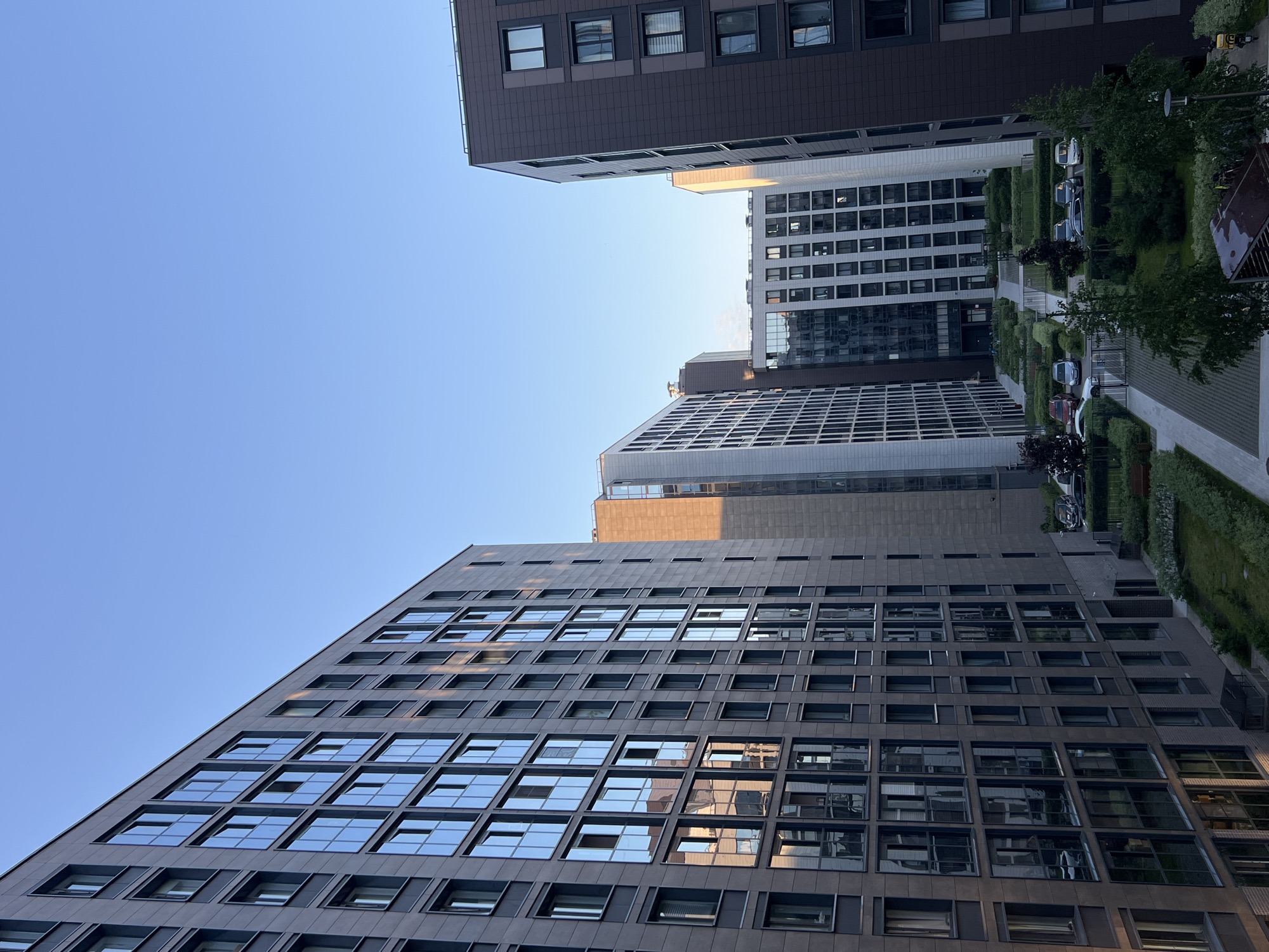 This image, rotated vertically, depicts a modern urban complex in Saint Petersburg, Russia, characterized by several tall, contemporary buildings arranged around a landscaped courtyard. The architecture is sleek and geometric, with buildings primarily featuring dark-toned facades, possibly dark grey or brown, accented by numerous square and rectangular windows. Many windows are highly reflective, mirroring the clear blue sky and elements of the adjacent structures. One prominent building on the left side of the frame exhibits a distinct angular profile and a repetitive grid of reflective glass panels.
The interior courtyard between the buildings is meticulously landscaped, featuring paved walkways, well-maintained green lawns, and strategically placed trees and hedges. Several cars are parked along the pathways, suggesting the area serves a residential or commercial purpose. The sky, visible in the upper and left portions of the image, is a bright and clear blue, indicating sunny weather. The natural lighting suggests it is daytime, likely late morning or early afternoon, with sunlight creating reflections and highlighting textures on the buildings.
No specific activities or people are visible, lending a quiet and orderly atmosphere to the scene. There is no discernible text within the image. The overall impression is one of modern urban planning and design.
