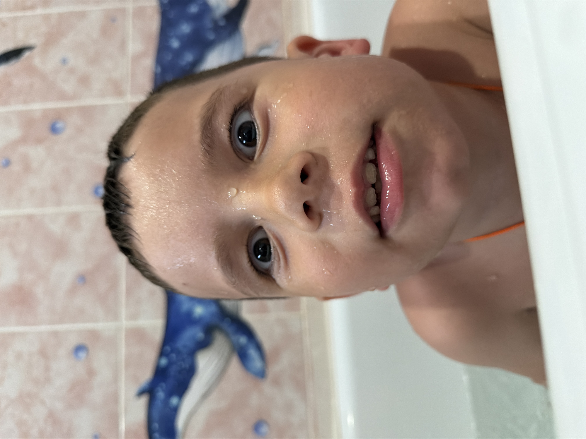 A young child with wet hair and face is sitting in a bathtub. The child is looking up at the camera. Behind the child is a tiled wall with a blue whale design. The location is a bathroom, possibly in an apartment or house. The city of Chaikovsky, Russia, is not discernible from the visual information.