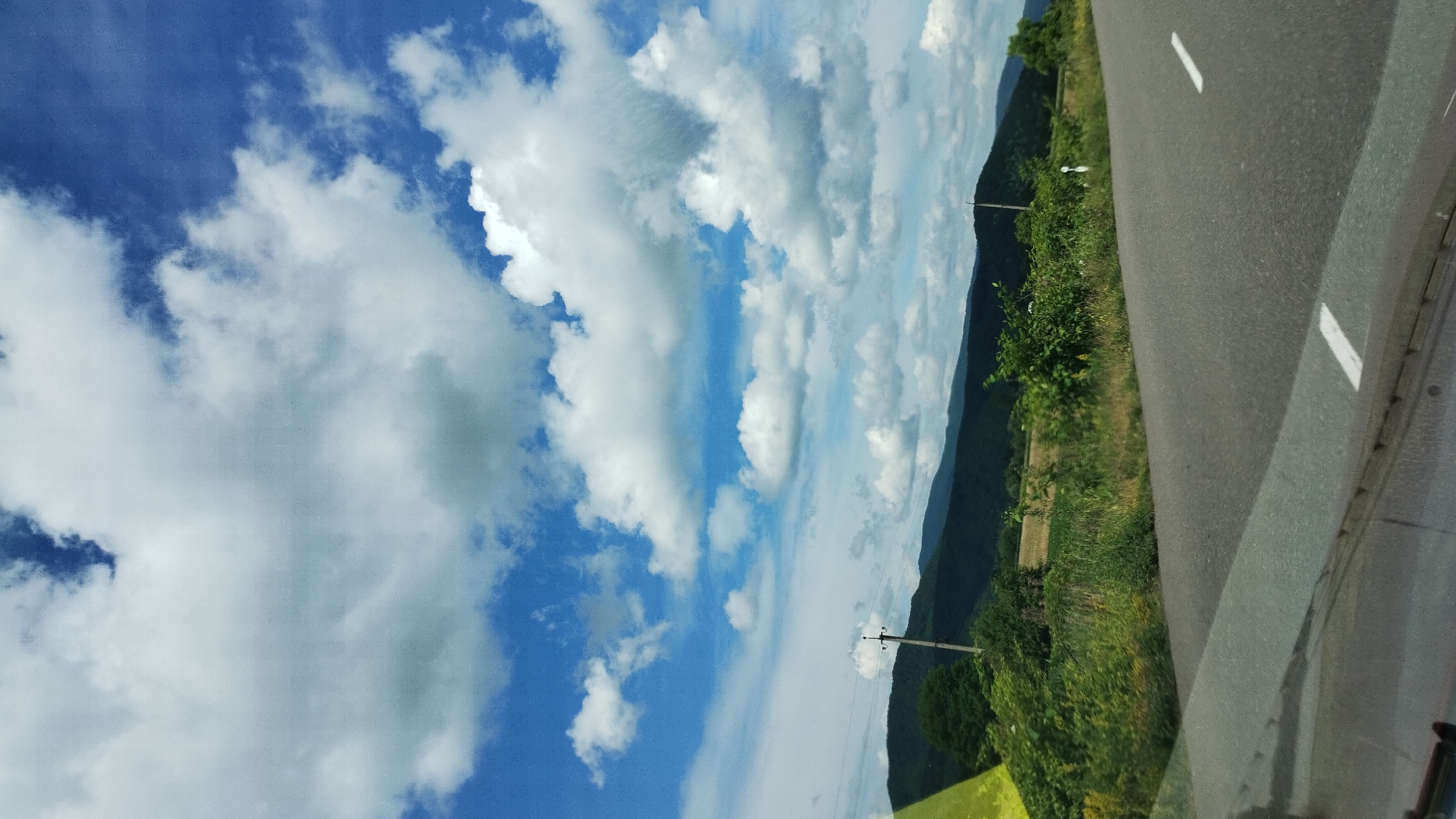 This is a landscape photograph taken from a vehicle, likely in Veliatyno, Ukraine. The image is oriented vertically.
The main subjects are the sky, a road, and a natural landscape. The sky is a prominent feature, filled with a mix of bright white and darker gray clouds against a vibrant blue backdrop. Sunlight suggests it's daytime. The road, with white lane markings, occupies the right side of the frame, indicating movement. To the left of the road, there is a lush green slope covered in trees and vegetation, with a glimpse of fields further down. In the distance, rolling hills or mountains are visible, their forms softened by the haze. A solitary utility pole with wires is present on the left side of the image.
No people are visible in the frame. The activity taking place is implied to be travel, as suggested by the road and the perspective from a moving vehicle.
Notable details include the dramatic cloud formations, suggesting potentially changing weather or a picturesque sky. The greenery points to a temperate climate, likely summer or late spring. The presence of the road and utility pole are common elements of rural or semi-rural landscapes.
There is no visible text in the image.