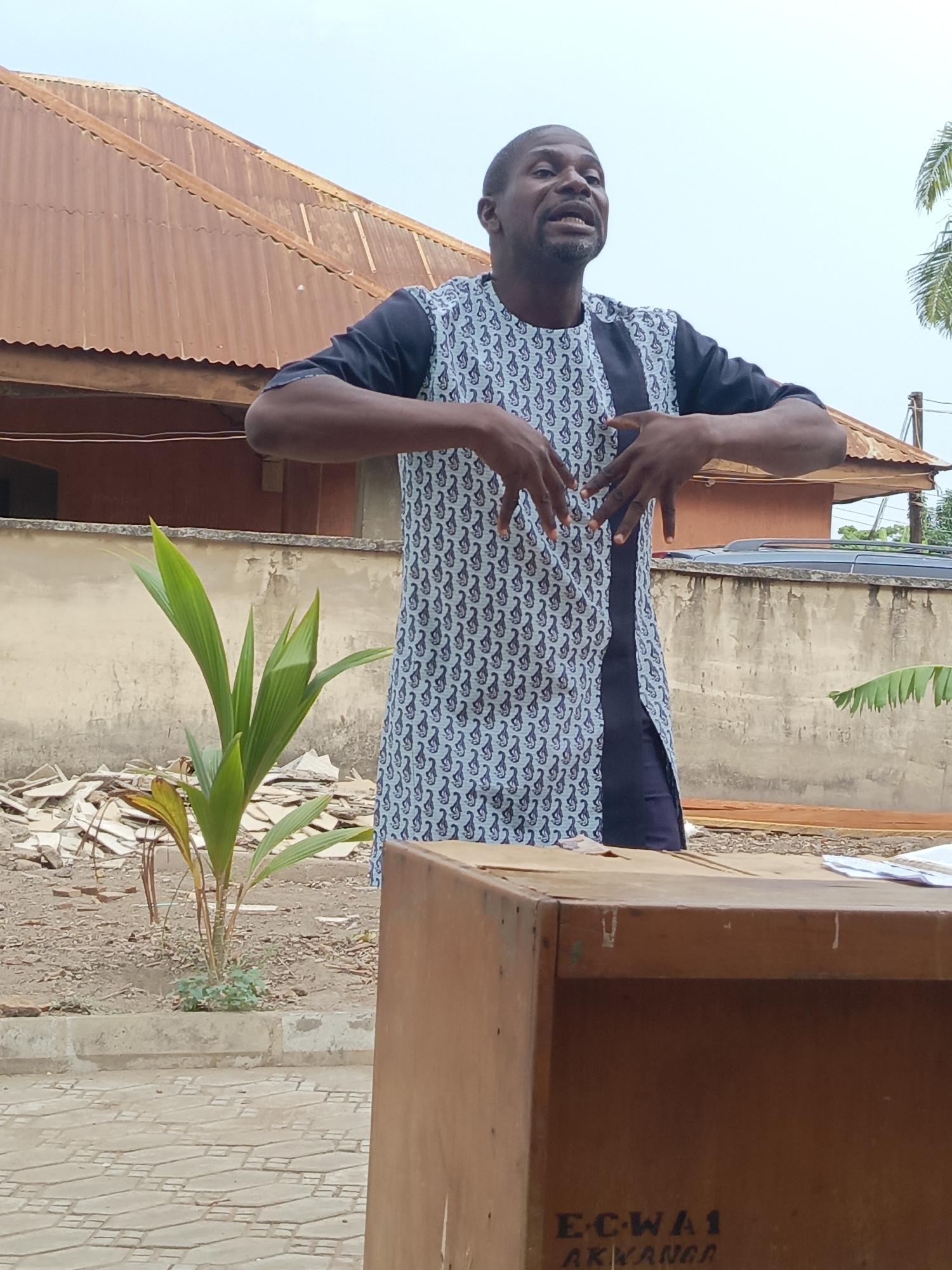 An outdoor, eye-level shot captures an adult male passionately speaking or presenting in what appears to be a courtyard or informal gathering space in Akwanga, Nigeria. The man, with dark skin, a beard, and short hair, stands centrally, wearing a long-sleeved tunic with a dark blue base and a light blue/grey patterned design, featuring a solid dark panel down the center. His mouth is open as if mid-sentence, and his hands are raised with fingers spread, conveying animated gestures. He looks slightly upwards and to the right, suggesting he is addressing an unseen audience.
The background features a clear, bright sky, indicating daytime, likely late morning or early afternoon, with no visible harsh shadows. To the left, a building with a rusty, corrugated metal roof is visible. A young, vibrant green plant (possibly a palm or coconut seedling) grows from the dirt ground to the man's left, surrounded by some scattered debris. Further back, a light-colored wall runs horizontally, behind which a dark vehicle's roof is partially seen on the right. To the far right, some green palm-like foliage is also visible. The ground in the foreground consists of patterned paving stones.
In the bottom right corner, a sturdy, light brown wooden box or lectern is partially visible. On its front surface, dark text is printed, which reads: "E-C-WA1 AKWANGA". A few white papers rest on top of this box. The man's expressive demeanor and energetic posture suggest he is deeply engaged in his speech, possibly delivering a sermon, lecture, or a public address to a group.