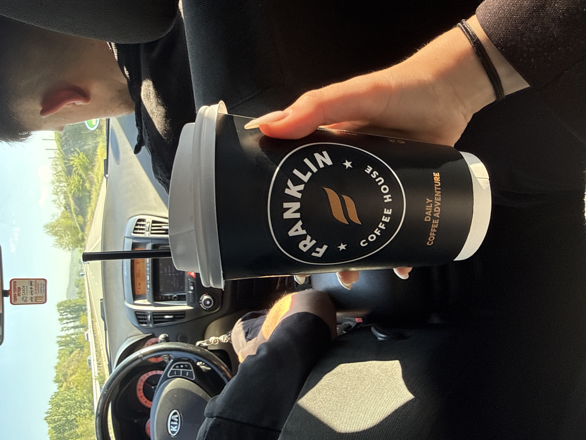 The image shows the interior of a Kia car driving on a road near Blajievo, Bulgaria. The steering wheel and dashboard are visible. A hand is holding a black Franklin House Coffee cup with a white lid and a straw. The driver is wearing black clothing and a bracelet. The rearview mirror has a tag on it. The landscape outside the window includes green trees and a blue sky. The road appears to be a highway.