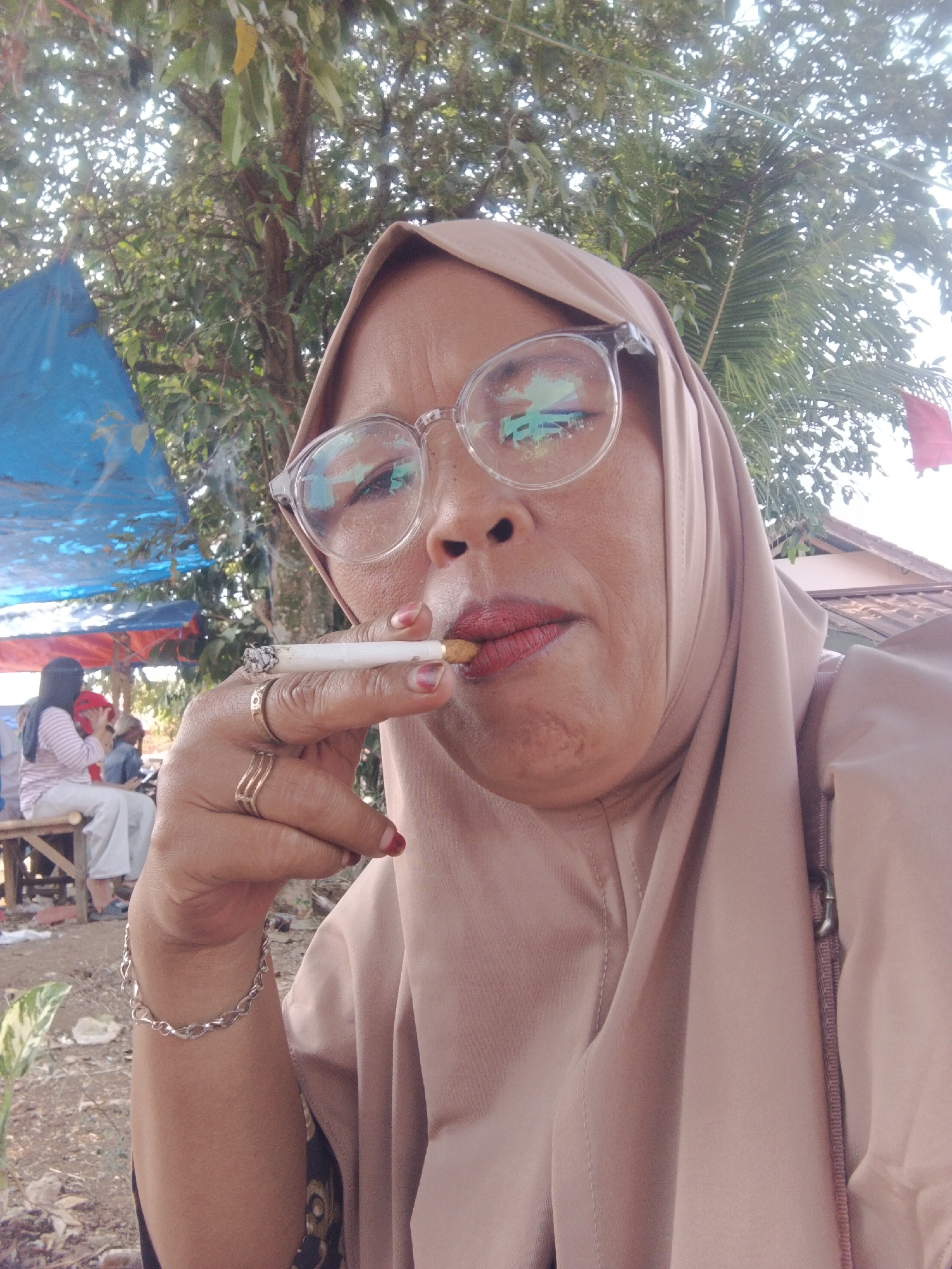 A middle-aged woman wearing a beige hijab and round eyeglasses is captured in a close-up, outdoor, selfie-style photograph. She is smoking a cigarette, with smoke visibly rising. Her lips are painted with red lipstick, and she wears two gold rings on her right ring finger, a silver bracelet on her left wrist, and has red nail polish on her fingernails. In the background, a blue and red tarp is visible, along with a tree and a glimpse of a building. Further in the background, two individuals are seated on a wooden bench, with one wearing a striped shirt and white pants, and the other a dark shirt. The setting appears to be a public area, possibly a market or street side, in Warungkiara, Indonesia.