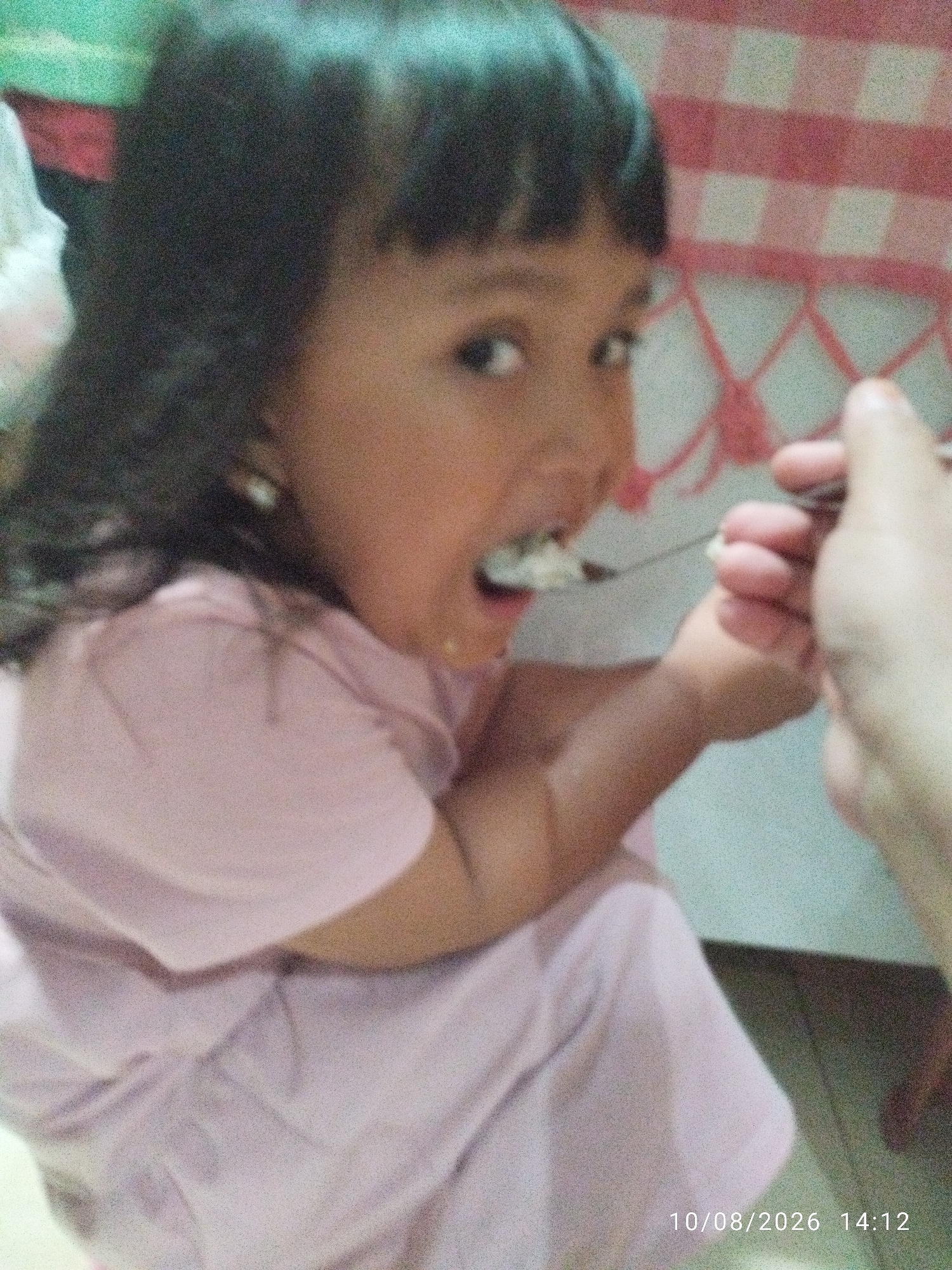 The image shows a close-up, overhead shot of a young girl with dark hair being fed rice from a spoon by an adult. The girl is wearing a pink top and has her mouth open to receive the food. The background features a red and white checkered pattern, likely a tablecloth, and a green surface. The overall lighting is dim and grainy, suggesting it was taken indoors with poor lighting conditions. The timestamp "10/08/2026 14:12" is visible in the bottom right corner. No specific location in Bandar Lampung, Indonesia can be identified from the image.