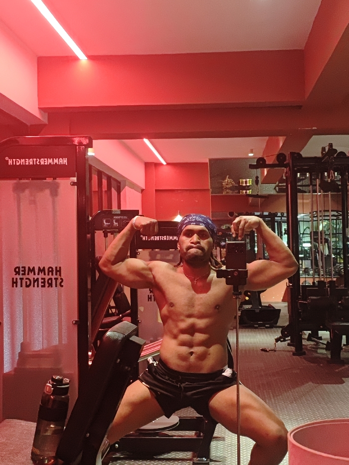 The image depicts a shirtless, muscular male posing in a gym located in Chongqing High-tech Zone, China. The environment is illuminated by red linear LED strip lights, casting a dominant red hue over the red-painted walls and ceiling. The male, central to the frame, wears black athletic shorts and a blue bandana over his head. He is actively flexing both biceps, exhibiting a well-defined abdominal region. He has facial hair, dark dreadlocks, a thin chain with a cross pendant, and a stud piercing on his left nipple. His expression is serious. He is taking a mirror selfie using a smartphone mounted on a small, dark tripod positioned in front of his waist. The mirror's reflection reveals additional gym equipment, including a black Hammer Strength machine to the left of the subject, displaying its logo in reverse. Further weightlifting apparatuses are visible to the right. A dark-colored water bottle stands on the textured floor near the base of the Hammer Strength machine.