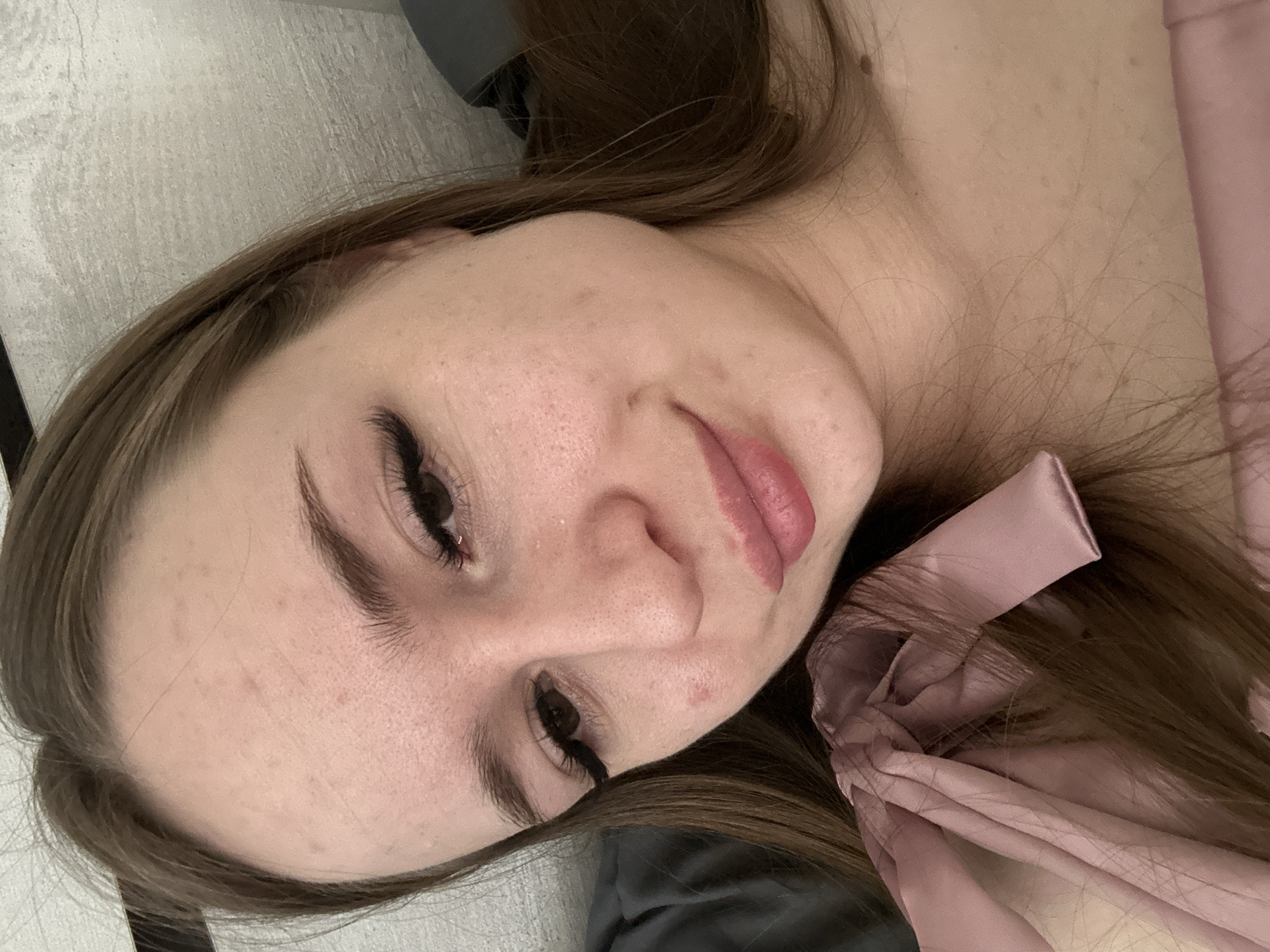 A close-up, rotated image shows a woman's face and upper chest. She is lying on a light-colored surface. Her hair is long and brown. She is wearing a light pink garment with a bow on the right side of the frame. Her eyes are open, and she has long, dark eyelashes. Her skin has some blemishes. The location is Unknown, Unknown.