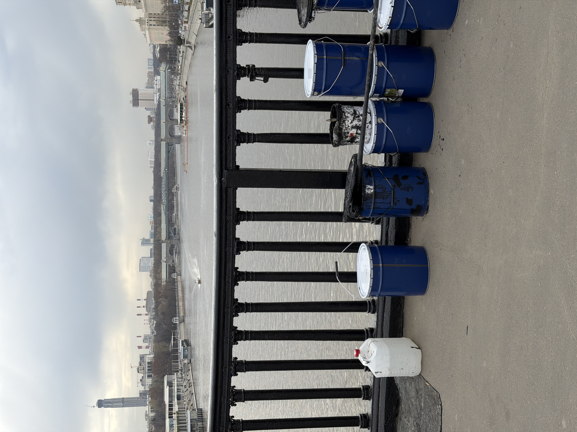 The photograph appears to be taken from a bridge in Moscow, Russia, likely the Bolshoy Kamenny Bridge, as the Kremlin is faintly visible in the background. The image is characterized by paint-related materials lined along the bridge's railing.
Subjects/Objects:
* Several blue paint buckets with white lids are aligned on the bridge next to the railing. Some show signs of usage with paint residue.
* There's also a white plastic container with a red cap sitting to the side of the buckets.
* The railing of the bridge is black, with decorative elements.
* The water of the Moskva River is visible, with at least one boat.
* In the background, the city skyline, with notable buildings including the Kremlin.
* The sky is overcast.
Actions/Interactions:
* The alignment of the paint buckets suggests they might be related to recent or ongoing bridge maintenance or painting.
* A boat on the river is navigating the water.
Environment:
* The weather appears overcast.
* The river seems calm.
* The location is urban, with buildings visible in the background.
Overall: The scene captures a moment of activity or rest during some maintenance or painting work on a bridge in Moscow. The composition of the image emphasizes the industrial or maintenance aspect against the backdrop of the city.