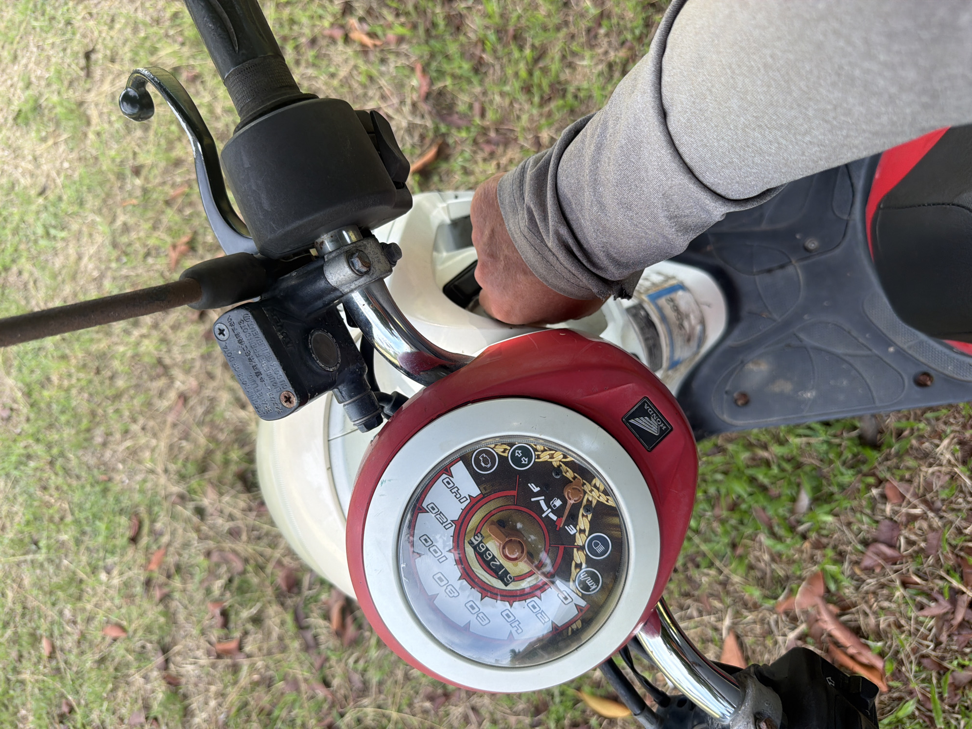 This is a close-up, high-angle, eye-level shot of a person's right hand operating the ignition of a red and white Honda scooter. The scooter's dashboard is visible, featuring a speedometer with a reading of "612666" km/h, fuel gauge indicating 'F', and various indicator lights. The scooter is parked outdoors on a grassy area with scattered fallen leaves, characteristic of a rural or semi-rural setting like Baan Mae Nam, Thailand. The focus is on the scooter's controls and the hand's action.