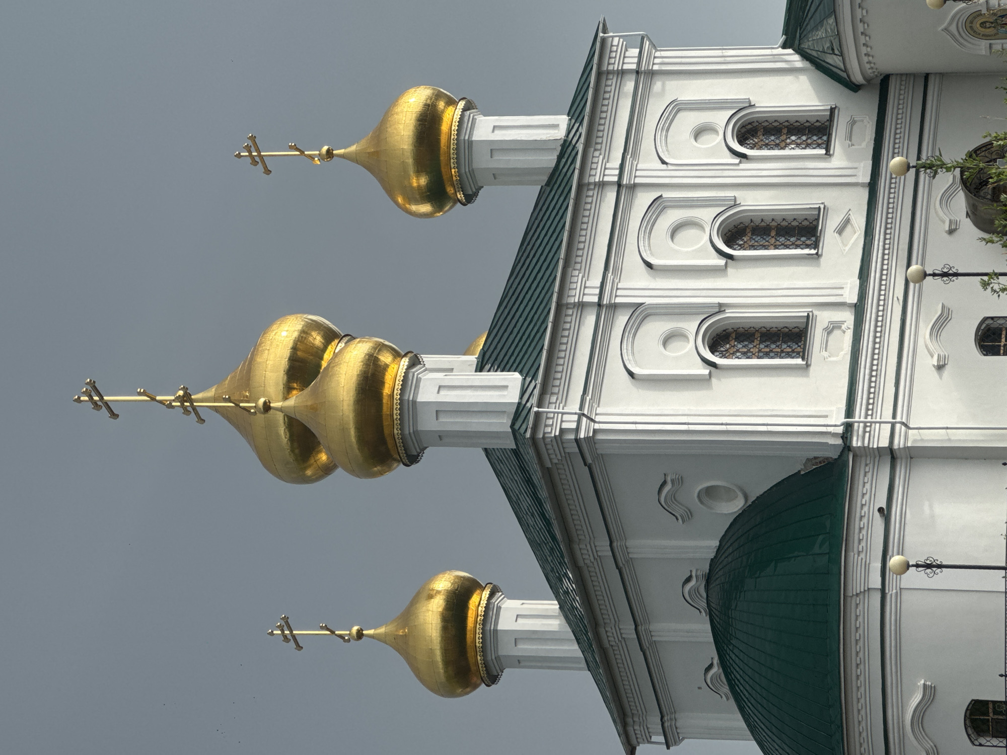 The image shows the exterior of a white building with multiple golden domes topped with crosses. The building has a green roof and features arched windows with decorative grates. The architecture suggests a religious building, likely a church, in Russia. The sky is overcast and gray, indicating cloudy weather. The image is oriented sideways, with the building appearing to extend upwards from the bottom of the frame. The composition focuses on the architectural details of the building, particularly the domes and the intricate window designs. The lighting is diffused due to the cloudy sky, creating a soft illumination of the structure. There is no visible text in the image. The scene appears to be set in a location with traditional Russian Orthodox architecture.