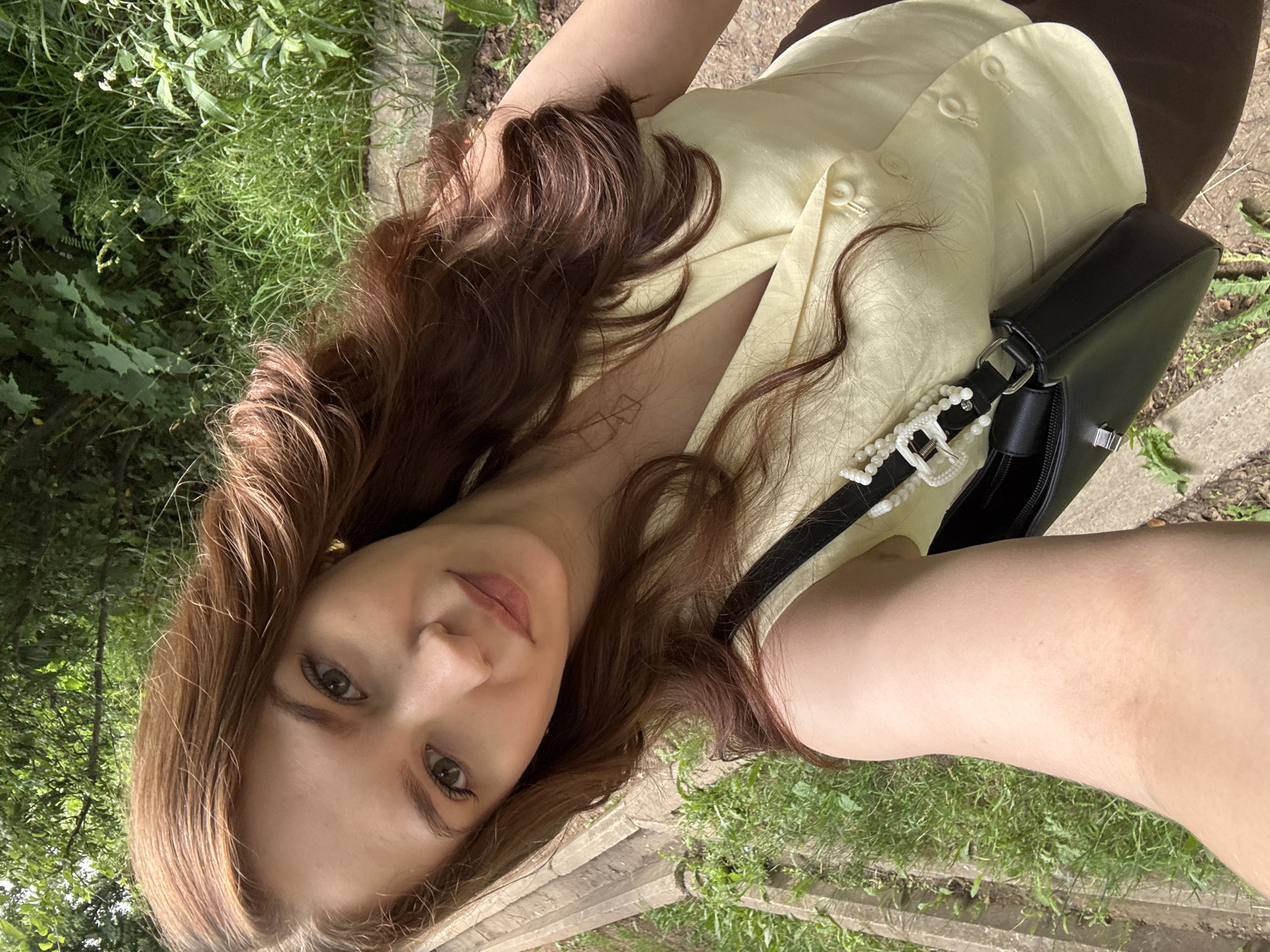 A young woman with long, wavy brown hair is taking a selfie outdoors. She is wearing a light yellow top with pearl buttons and a dark skirt or trousers. A black handbag with pearl detailing is slung over her shoulder. The background is lush green foliage and what appears to be a pathway or steps. The lighting suggests it is daytime, possibly sunny, given the shadows. The woman's expression is neutral, and the overall tone of the image is casual and relaxed. There is no visible text in the image.
