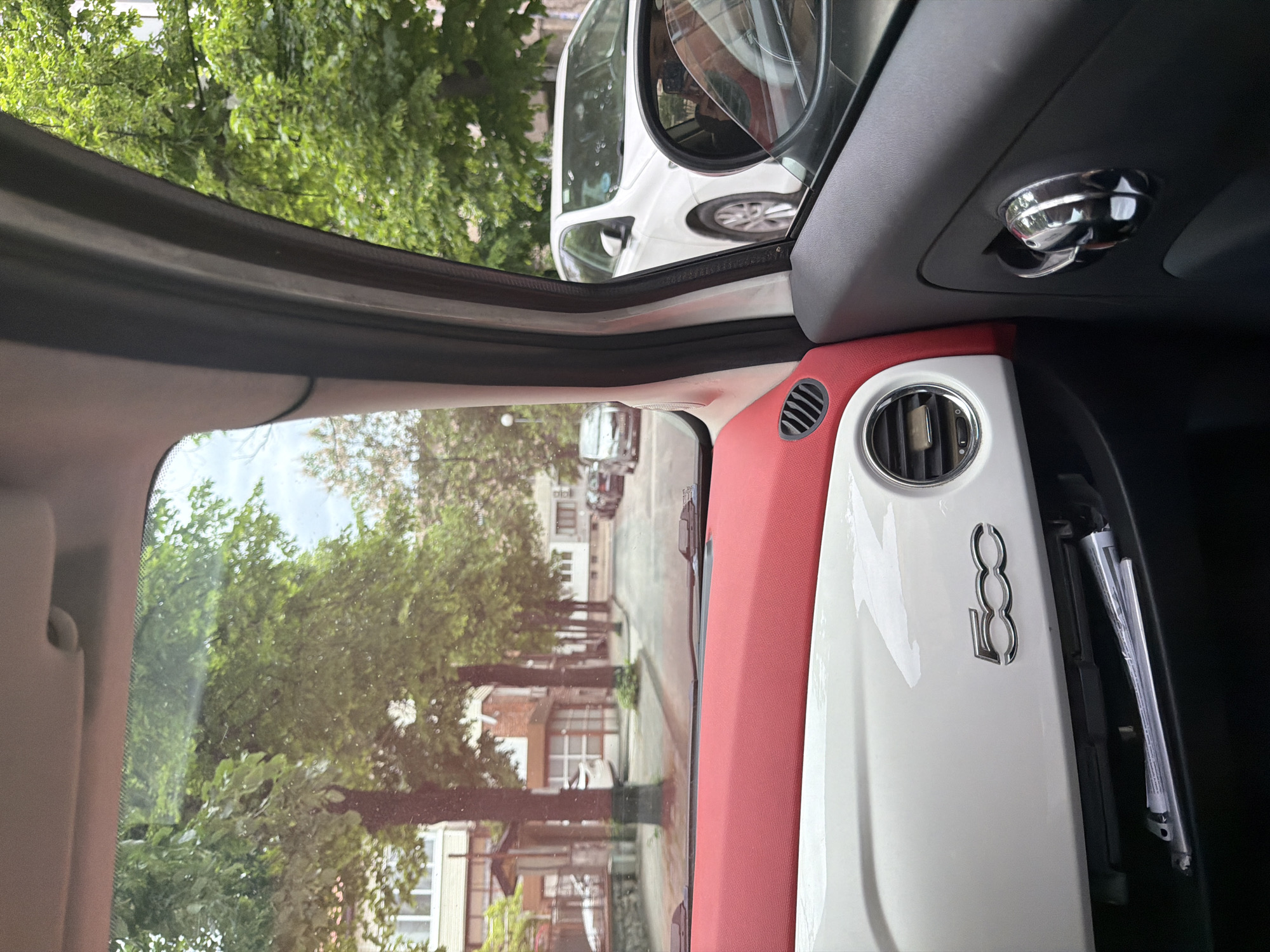 The image is taken from the interior of a white and red Fiat 500, with the door open. The interior trim is red and white. A chrome Fiat 500 logo is visible on the dashboard. The interior also features a chrome air vent and a silver dome-shaped fixture on the ceiling. Through the open door, a street scene is visible with trees lining the sidewalk, buildings, and other parked cars. The location is in a city, but the specific city is unknown.