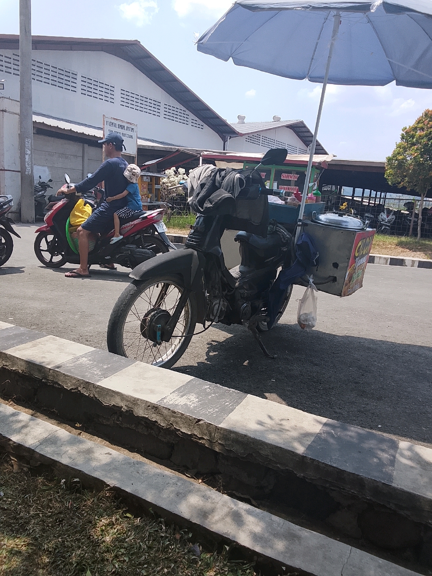 In Karangpucung, Indonesia, a father and child are riding a red motorcycle. The father, wearing a dark blue long-sleeved shirt and dark shorts, is driving. A child, in a blue shirt and striped shorts, is sitting in front of him, holding on. The motorcycle is equipped with a large, light blue umbrella for shade. Attached to the back of the motorcycle is a cart, likely for selling goods, with a large silver pot and a clear plastic bag hanging from it. In the background, industrial-style buildings with white walls and slatted vents are visible under a bright, partly cloudy sky. Other motorcycles are parked or in motion nearby. The scene takes place on a paved road next to a concrete divider and a grassy area.