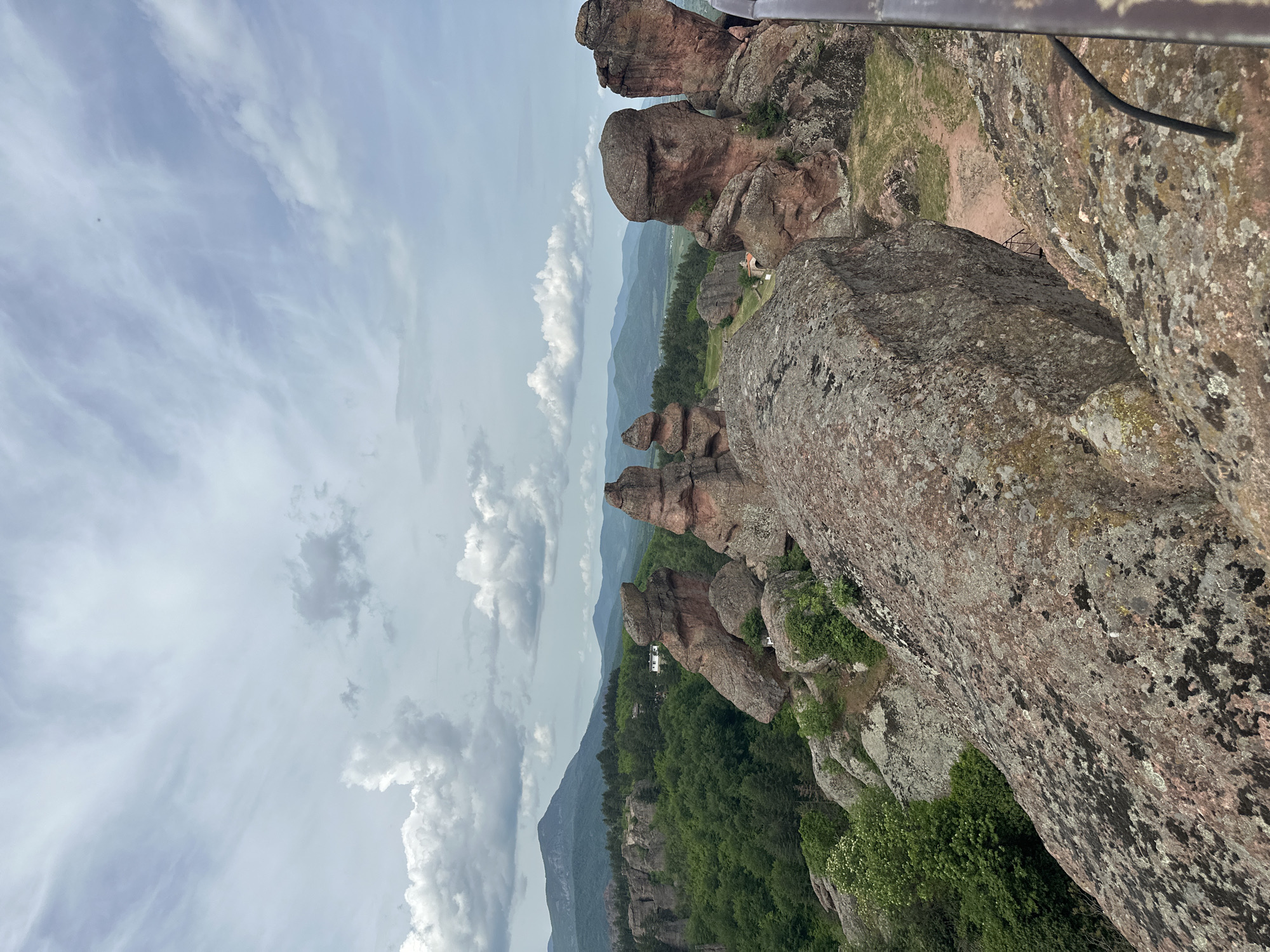 The image depicts the Belogradchik Rocks in Belogradchik, Bulgaria. The prominent rock formations are composed of reddish sandstone and conglomerate, rising above a landscape of green forest and rolling hills. The sky is filled with light and dark clouds. A part of a metallic safety railing is seen at the top. The rock formations stretch into the distance, with visible mountain ranges in the background. The scene is captured from a high vantage point, giving a panoramic view.