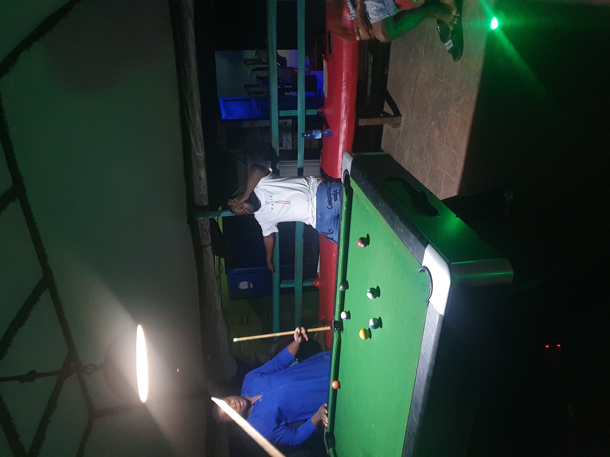 The image depicts an indoor billiards game occurring at night in Lokoja, Nigeria. A person in a blue shirt is positioned at the left side of a green pool table, holding a pool cue and preparing to strike a ball. Another person, wearing a white "PARIS" t-shirt and denim shorts, is seated on a red bench in the background, gesturing with their hands above their head. Several billiard balls are scattered across the table, illuminated by an overhead light fixture. The environment appears to be a dimly lit recreational space with a wooden ceiling and green walls. There is a bottle of water on the bench and a blue cooler in the background. The floor is tiled. The overall atmosphere suggests a casual gathering or social event centered around the game of pool.