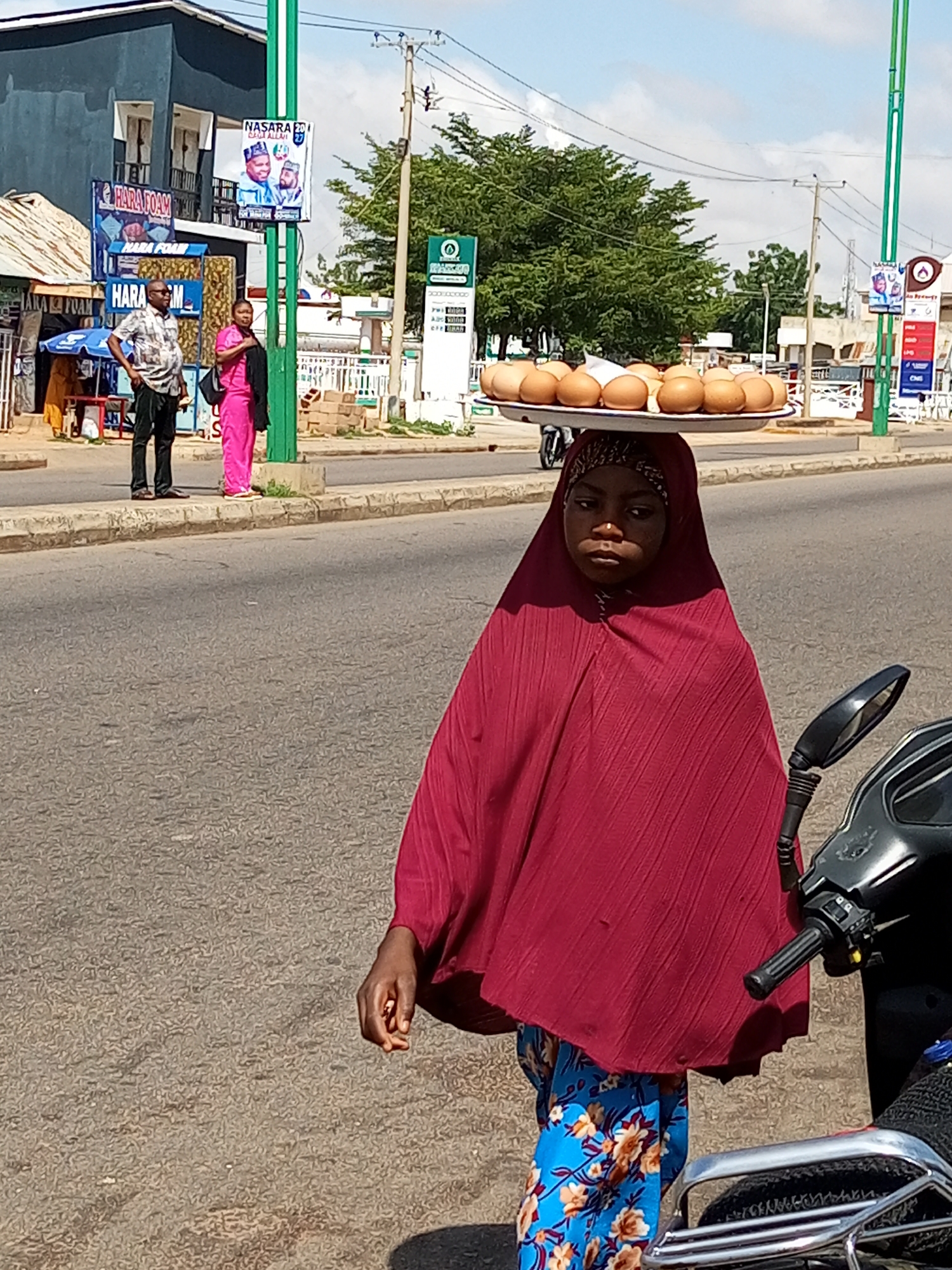 In Garko, Nigeria, a young girl, appearing to be of African descent, is captured in a medium shot as she walks along an asphalt road. She is wearing a vibrant maroon hijab and a blue skirt adorned with a floral pattern. Balanced precariously on her head is a large white tray filled with numerous brown eggs. Her expression is neutral, and her gaze is directed slightly downwards. To her right, a portion of a black motorcycle is visible, with its handlebars and side mirror in frame. In the background, a green pole with a white sign is situated near a building. Further back, trees and other structures are discernible, suggesting a bustling urban environment. Two adults, a man in a patterned shirt and dark pants and a woman in a bright pink outfit, are standing on the sidewalk in the mid-ground, to the left of the girl. The overall scene depicts a street view with elements of commerce and daily life.