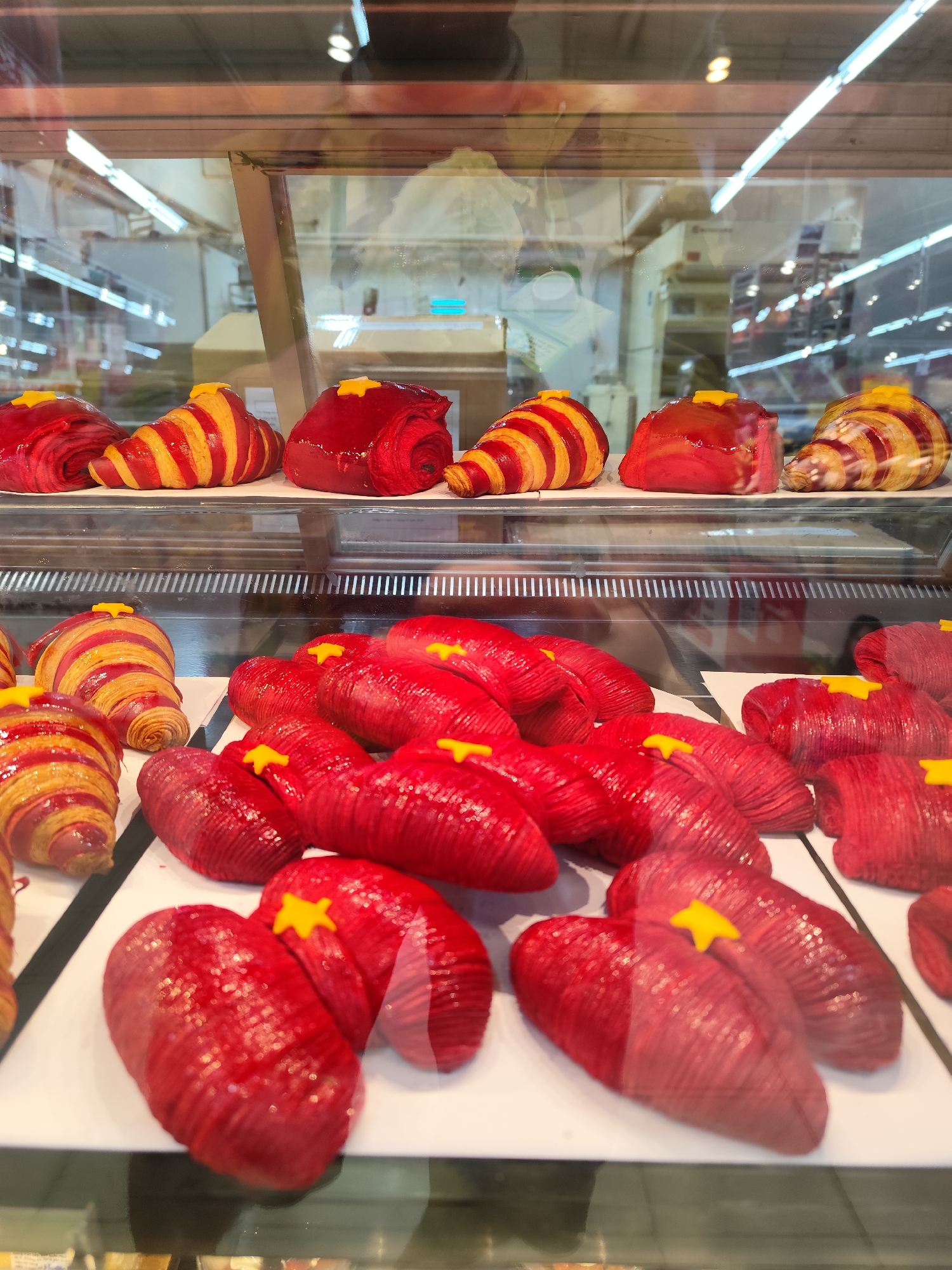 The image captures a bakery display case in Da Lat, Vietnam, filled with a variety of pastries. The pastries are predominantly red, with some featuring red and yellow stripes, and are topped with a yellow star. The display case itself is modern, with a glass front that reflects the overhead lighting and a glimpse of the bakery's interior. The pastries are neatly arranged on white trays, suggesting a clean and organized environment. The pastries seem to be creatively designed. The display showcases the pastries in an appetizing way. The background reflects bright lights, white walls, and bakery equipment.