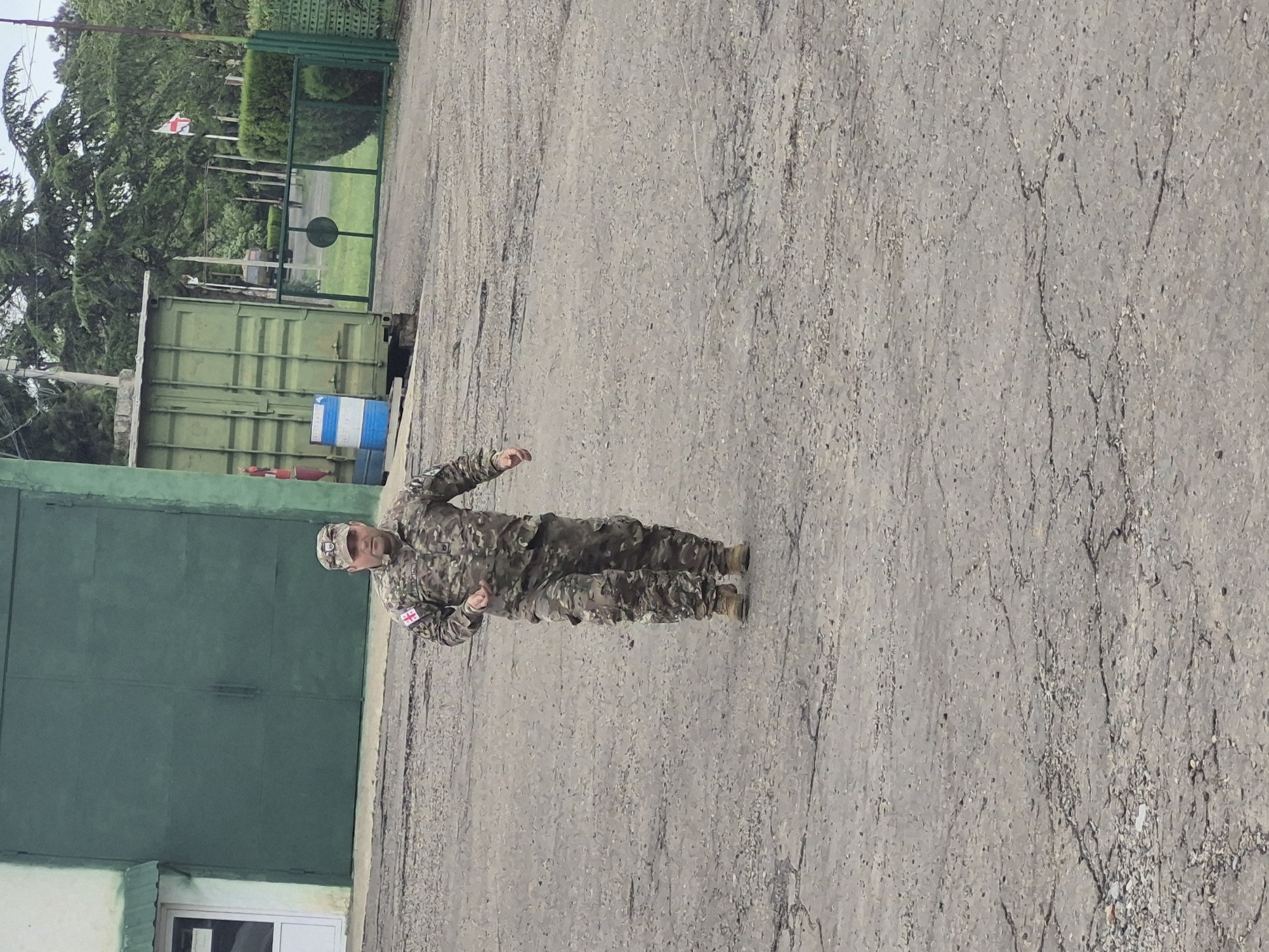 A man in military camouflage uniform stands on a cracked asphalt surface, facing the left of the frame. He is wearing a cap and uniform with a Georgian flag patch on his left shoulder. Behind him, there is a green corrugated metal building, with a partially visible white window frame on its lower left corner. Next to the building, there are a couple of blue and white striped barrels. In the background, there is a green metal gate and a Georgian flag waving in the wind. The image appears to be taken outside, with overcast lighting, and the asphalt surface shows signs of wear and damage. The specific location in Tbilisi, Georgia, cannot be determined from the image alone.