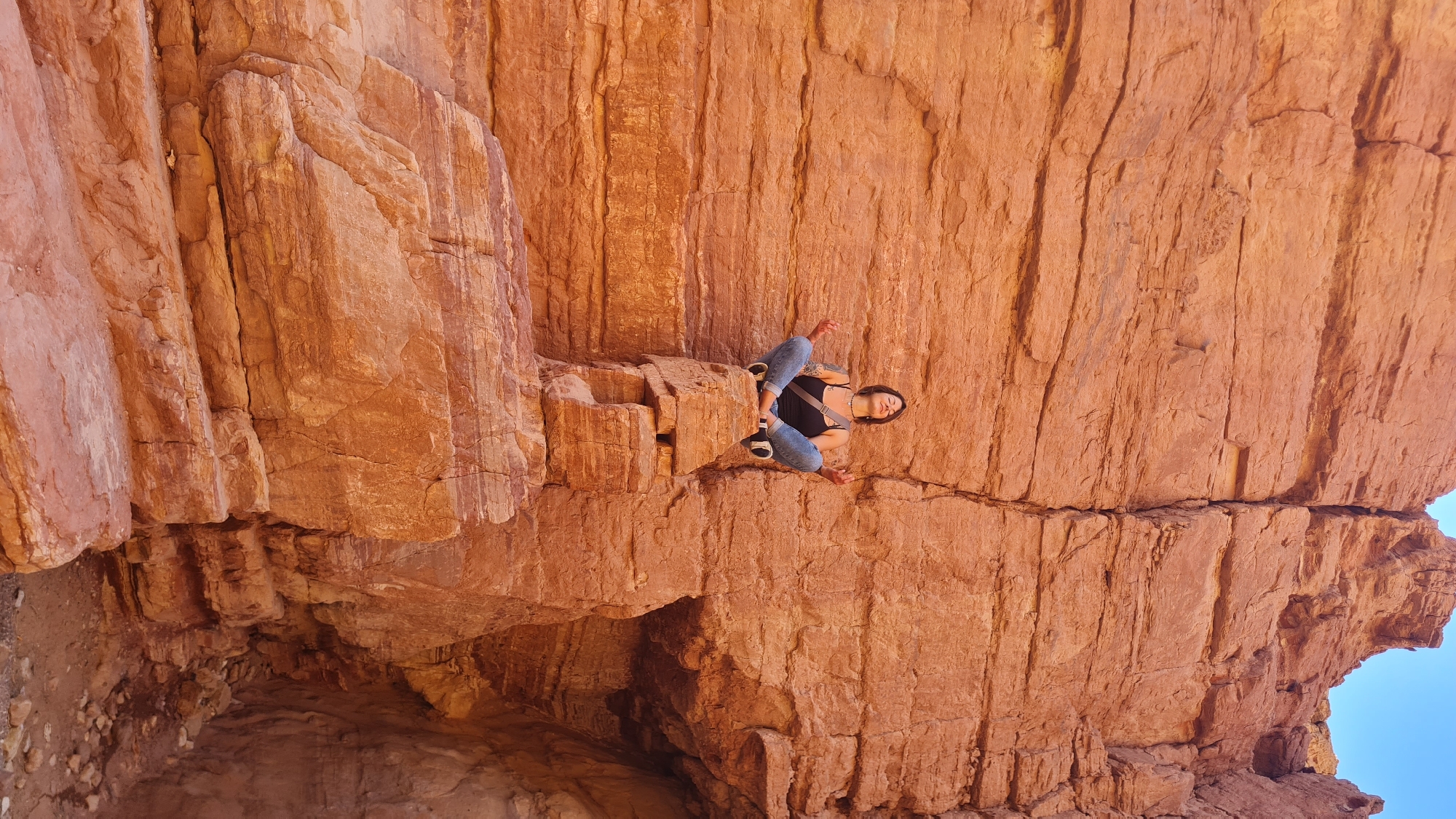 A medium shot shows a woman perched on a rock ledge within a deep, narrow slot canyon in Hevel Eilot Regional Council, Israel. The canyon walls are composed of reddish-brown rock with vertical striations and fractured surfaces. The woman is facing the camera and sitting with bent legs. She is wearing jeans, a dark tank top, and sneakers. A strap crosses her chest. The sky is visible in the upper right corner. The photograph is rotated.