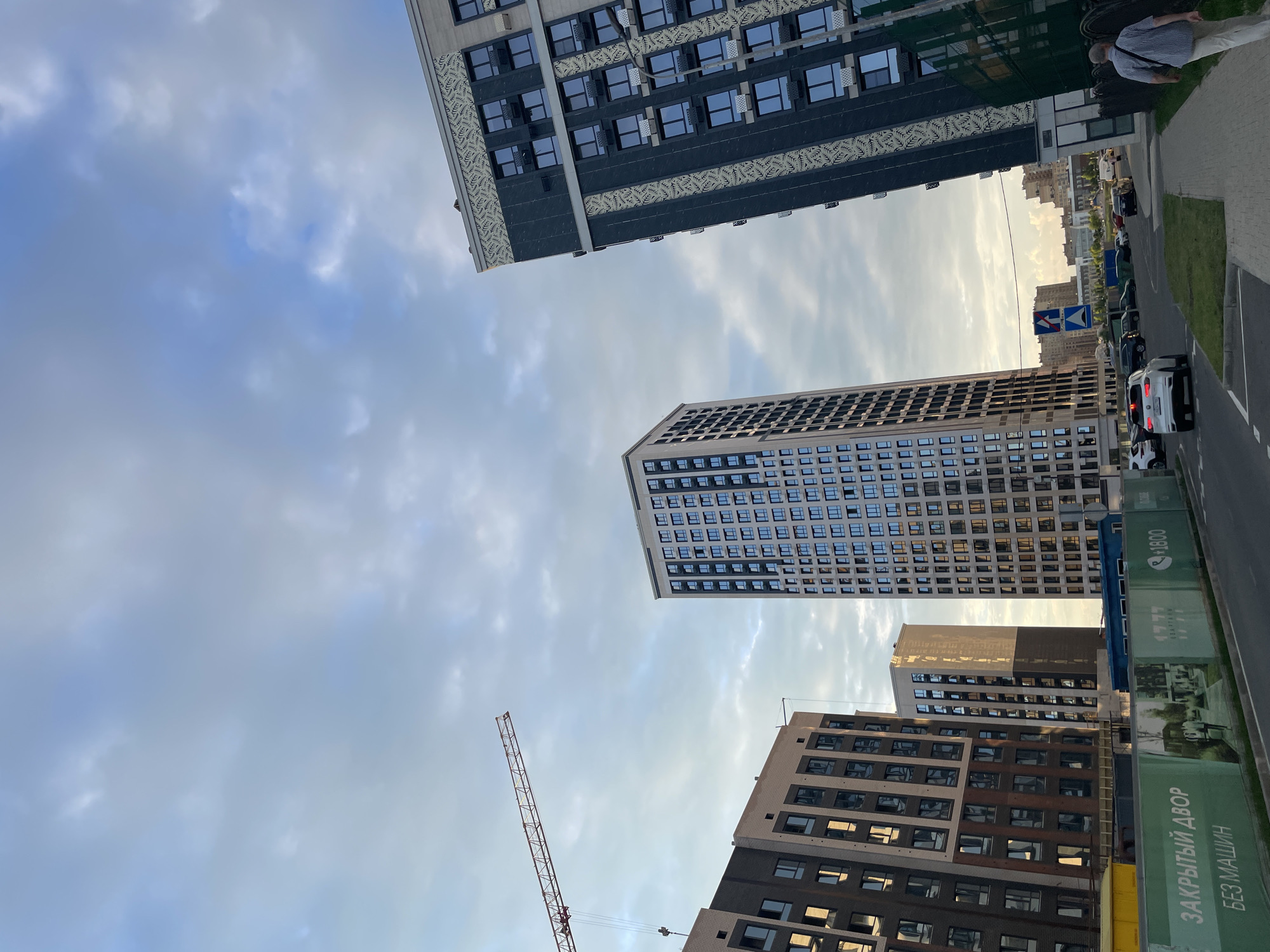 The image depicts a contemporary urban construction site in Stavropol, Russia, under a cloudy sky. In the foreground, a green fence with Russian text and images of residential buildings lines a paved road. Several modern, high-rise apartment buildings are visible, with one prominently featuring dark gray and white decorative elements. A red construction crane extends from the left side of the frame, indicating ongoing development. Cars are present on the road, and a person is walking on the sidewalk to the right of the frame. The overall scene suggests a developing residential area.