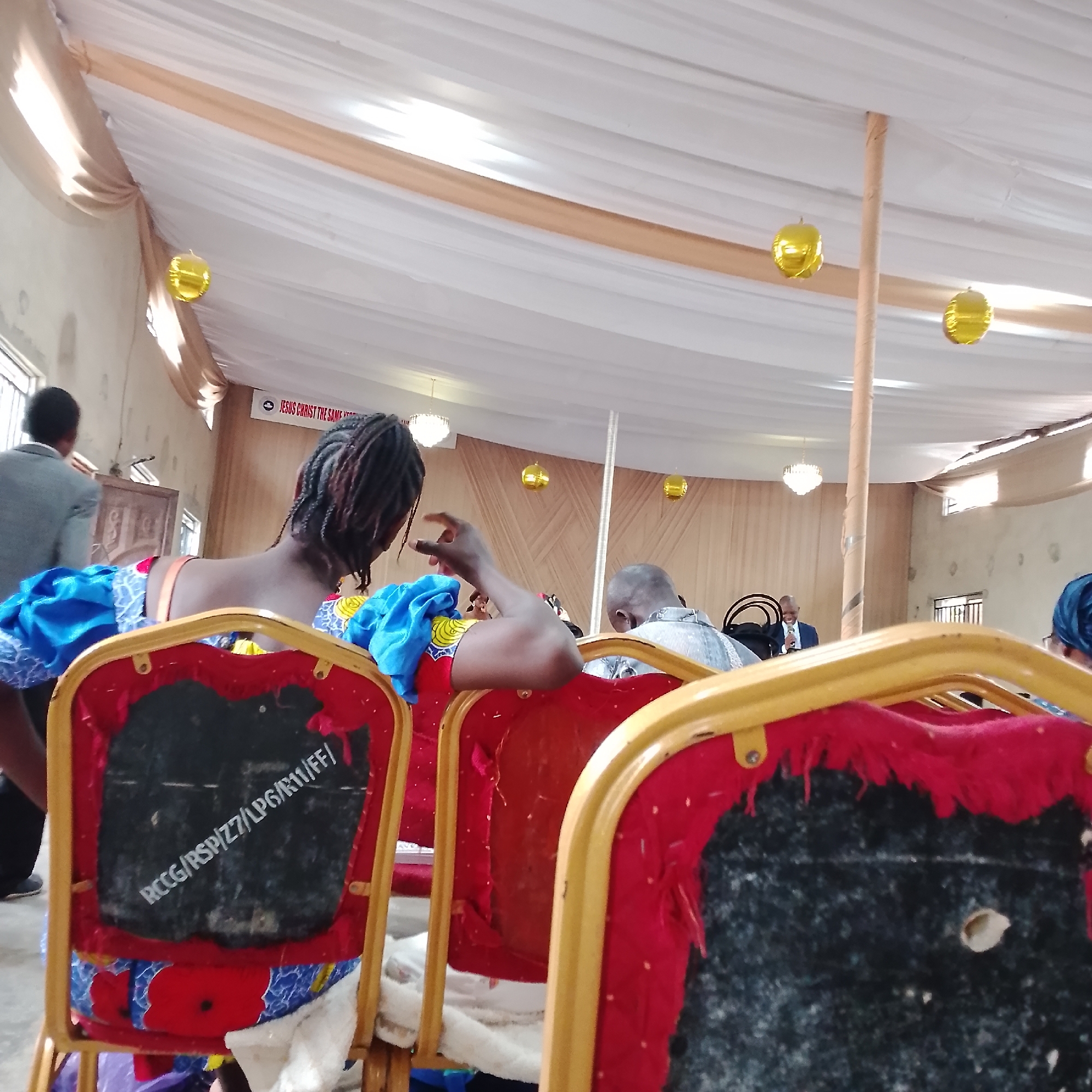 The image depicts an indoor gathering, likely a church service, in Ado, Nigeria. The ceiling is draped with white and beige fabric, adorned with gold balloons. A banner reading "JESUS CHRIST THE SAME YEAR" is visible on the wall. In the foreground, the backs of several red chairs with yellow frames are prominent. One chair has a black insert with white writing that appears to be an alphanumeric code, possibly "RCCG/RSP/Z7/LPG/R11/FF/". Several individuals are seated, facing away from the camera. A man in a grey suit is seen leaving the room on the left, and further back, a man in a light-colored shirt is visible. A woman with braided hair in a blue and yellow patterned dress is in the mid-ground. The overall scene suggests a religious event or meeting.