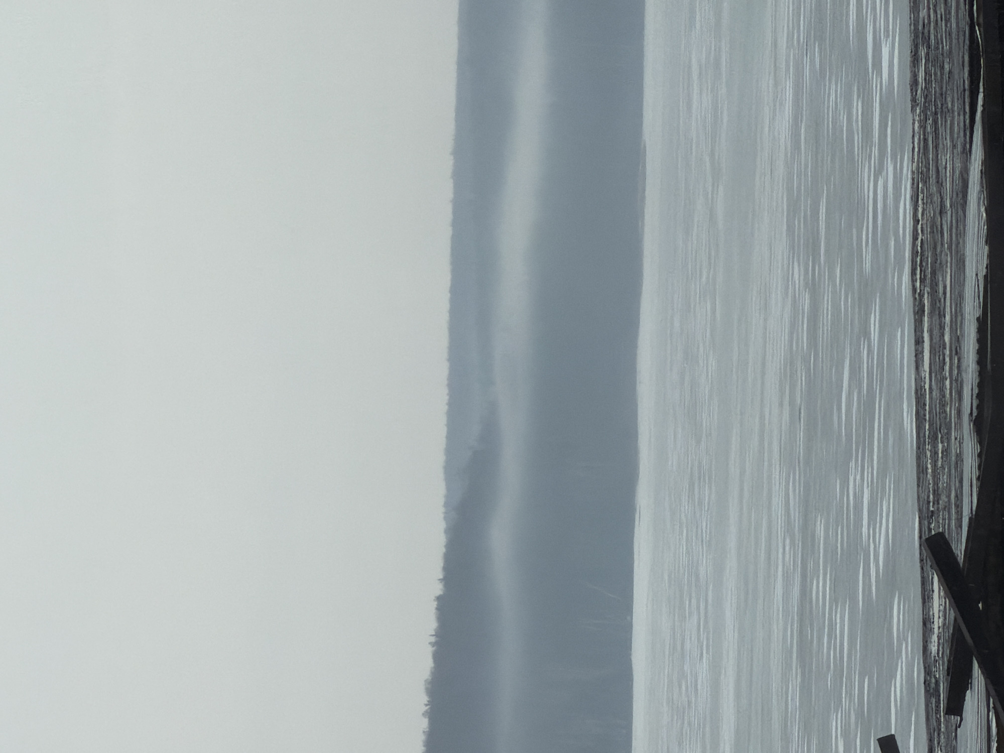 The image shows a waterfront scene in Chaikovsky, Russia, on a hazy or overcast day. In the foreground, there's what appears to be a wooden pier or dock structure extending into the water. The water itself is calm, with faint ripples suggesting slight movement. In the distance, there is a landmass across the body of water, likely the opposite shore of a river or lake. The hazy atmosphere obscures the details of the distant shore, but it's possible to discern the outline of trees or vegetation. The sky is overcast and uniformly grey, contributing to the overall subdued and muted tones of the scene. There is no visible activity on the water or along the shore.