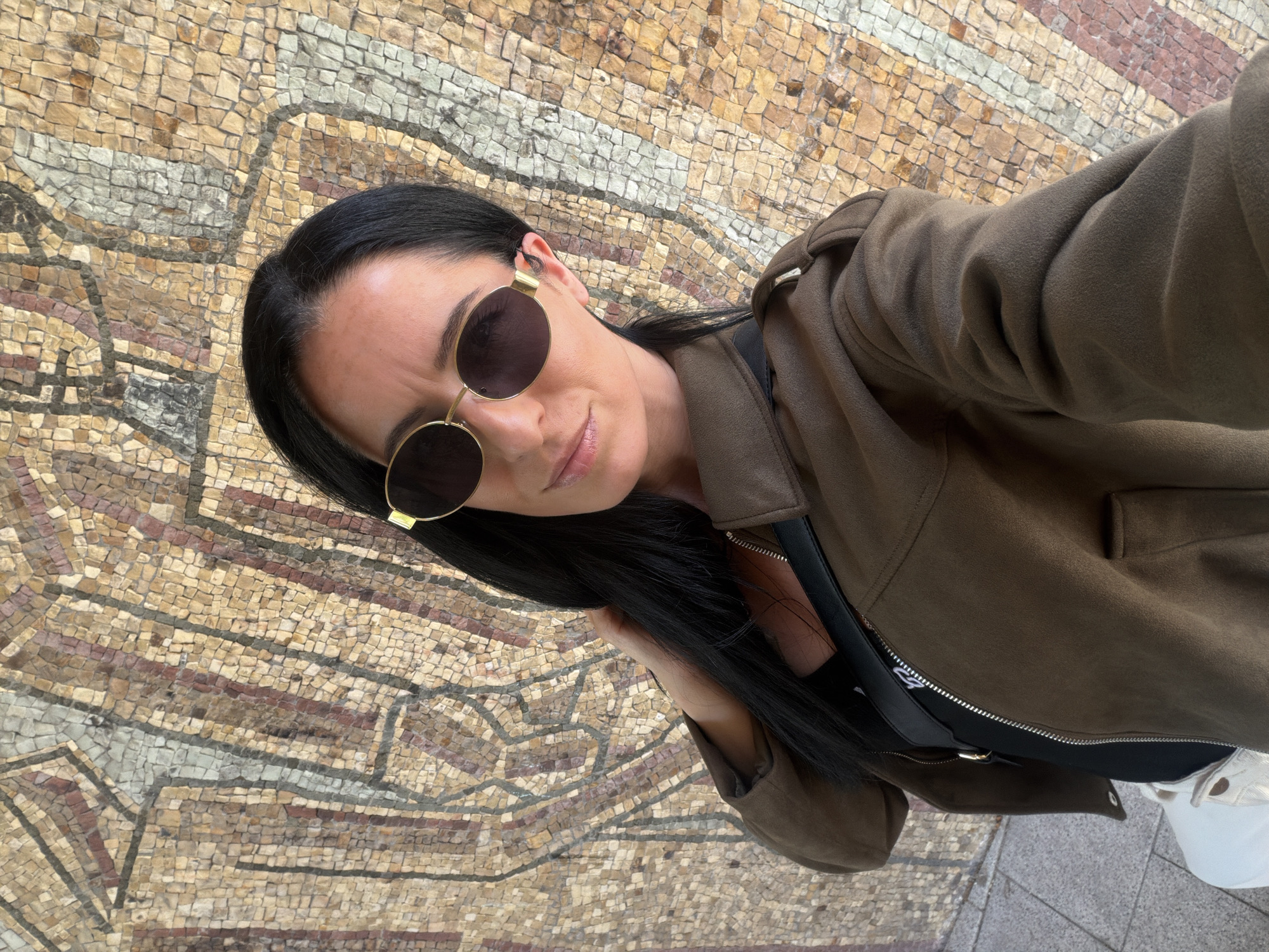 A woman with black hair and round sunglasses is taking a selfie in front of a mosaic wall. She is wearing a brown jacket over a black top and carrying a black purse. The mosaic is made of small, irregularly shaped tiles in various shades of brown, beige, and grey, forming an intricate pattern. The location is likely in Pleven, Bulgaria.