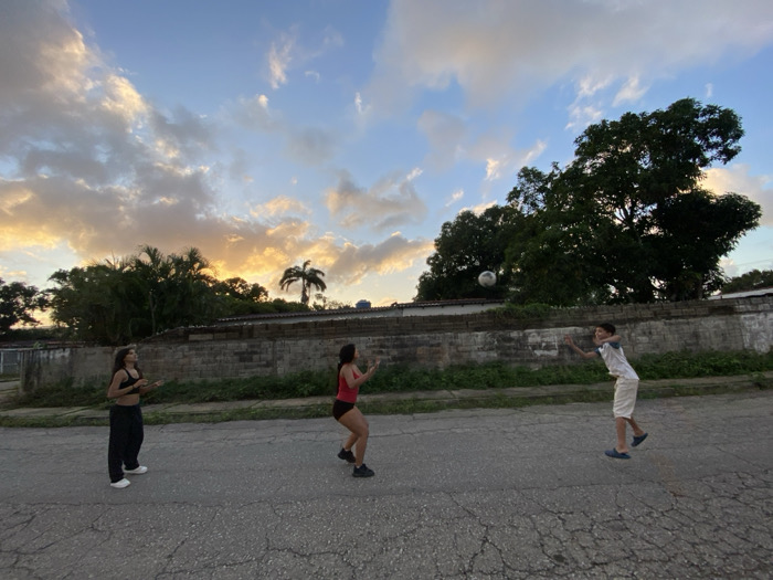 Three figures play in the middle of a cracked asphalt road. To the left is a person in black pants and a black top. In the middle, a figure is dressed in a red top and black shorts. To the right, a child is in mid-air, wearing a white t-shirt and white shorts. The trio appear to be playing with a ball. Behind them is a block wall, with various trees and foliage behind that. The sky is blue with golden clouds.