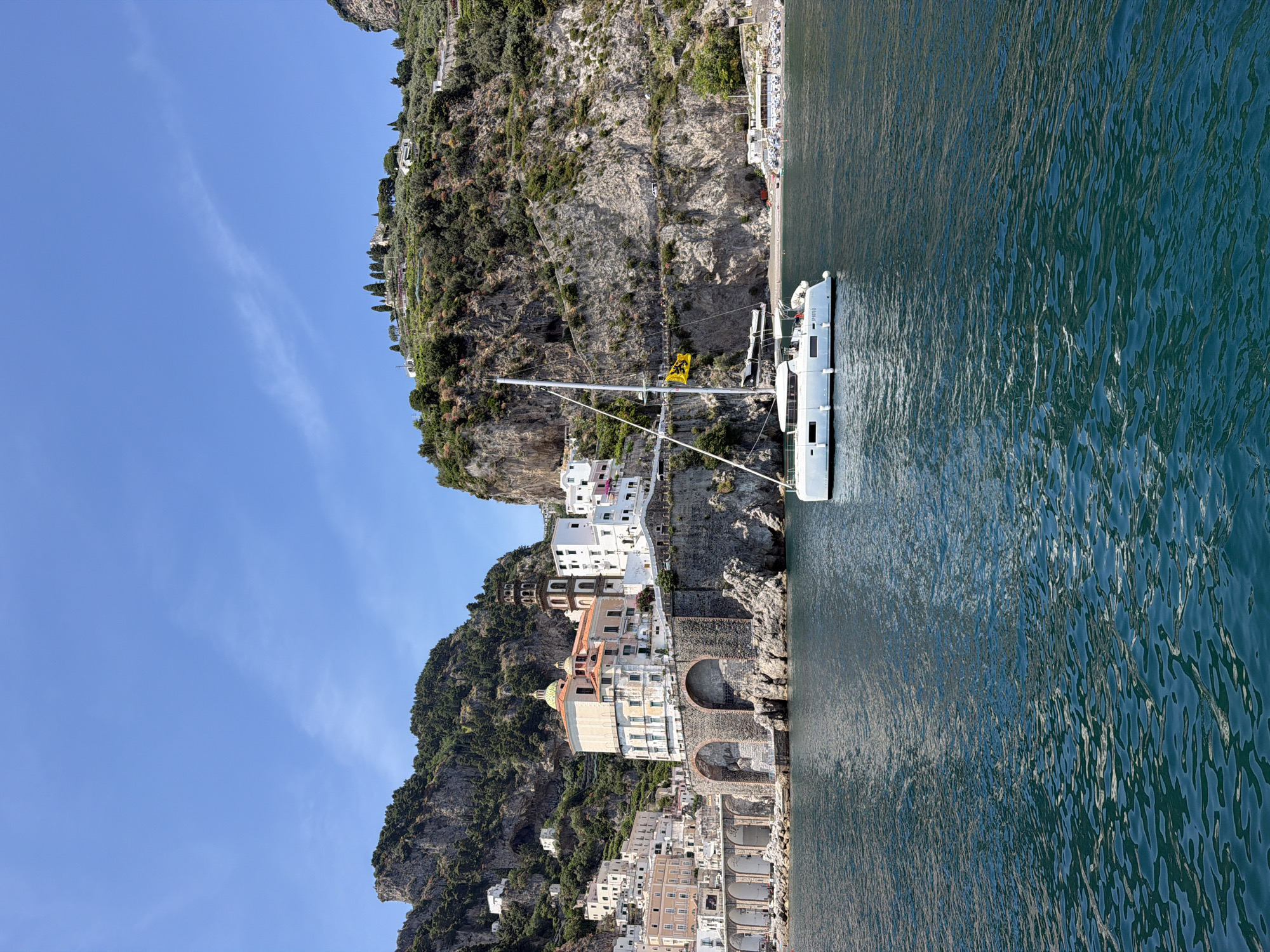 The image depicts a coastal scene in Ravello, Italy. A white sailboat is moored in the water near a steep, rocky coastline. The town is built into the cliffs, featuring white buildings with terra cotta roofs. Notable architectural features include arched structures along the waterfront and a domed building further up the slope. Vegetation covers the upper portions of the cliffs. The sky is blue with faint cloud formations. The water has a dark blue color. A beach can be seen.