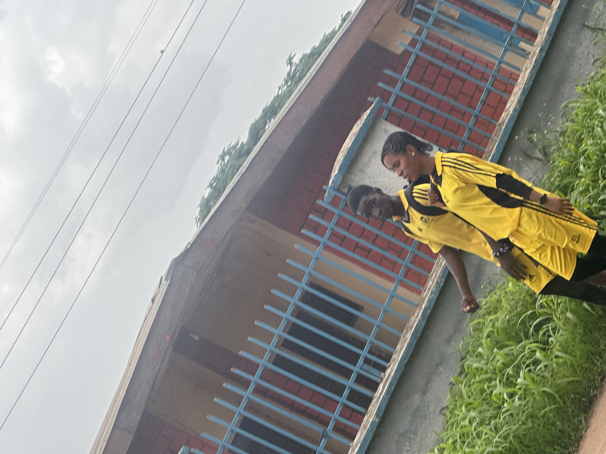 The image depicts two individuals, a young male and a young female, walking outdoors. They are both wearing matching yellow athletic uniforms with black accents. The male is on the left, wearing a short-sleeved shirt and shorts. The female is on the right, wearing a long-sleeved shirt with black under-sleeves and shorts. They are walking from left to right in the frame, with the male looking slightly to his left and the female looking forward and slightly up. Behind them, there is a building with red brick walls and a brownish-red roof, partially obscured by a blue metal fence. Green foliage is visible in the foreground, growing along a textured concrete barrier. The sky is overcast. The image appears to have been taken in an outdoor setting that could be a school campus or a public area in Gura Top, Nigeria.