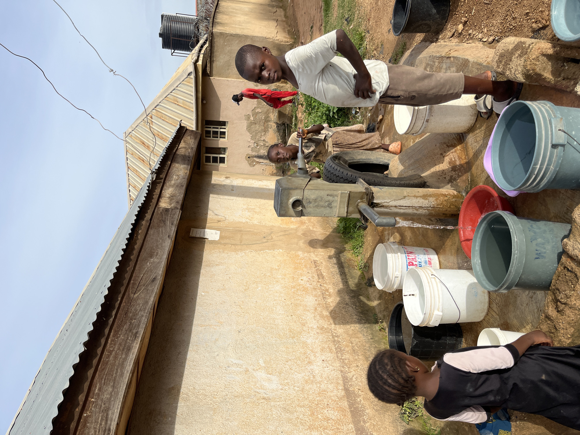 In Rafin Par, Nigeria, a scene unfolds at a water pump where four children are gathered. A young girl, facing away from the camera, stands to the left, her braided hair visible. She wears a black dress with light pink sleeves. Beside her, a large black bucket sits on the ground. To her right, a white bucket is filled with water, and behind it, another white bucket with red lettering is also full. Further right, a red basin contains water, and a large grey bucket is positioned in the foreground, filled with water and bearing faint blue lettering.
Behind these containers, two other children are actively engaged with the water pump. One child, wearing a light brown shirt and skirt, operates the pump, and a used tire is propped against it. The other child, a boy in a white t-shirt and brown shorts, stands looking towards the camera, his hands on his hips. In the background, a person in a red shirt and red pants walks away from the scene, and a large black water tank is visible atop a building. The environment is rustic, with a concrete wall of a building forming the backdrop, a corrugated metal roof extending over a portion of it, and sparse vegetation growing around the pump. The ground is a mix of dirt and concrete.