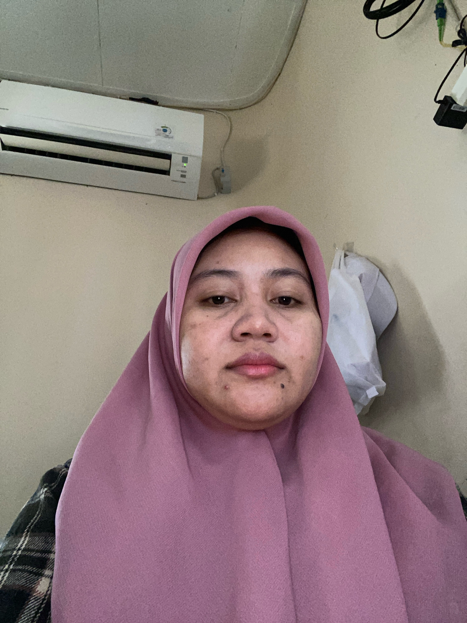 The image is a portrait of a woman wearing a pink hijab and a plaid shirt. She is looking directly at the camera. Behind her, an air conditioning unit is mounted on the wall. The background wall is a neutral color, with some electrical cords and a white bag hanging on a hook. The lighting appears to be indoor. The image quality is moderate with visible grain.