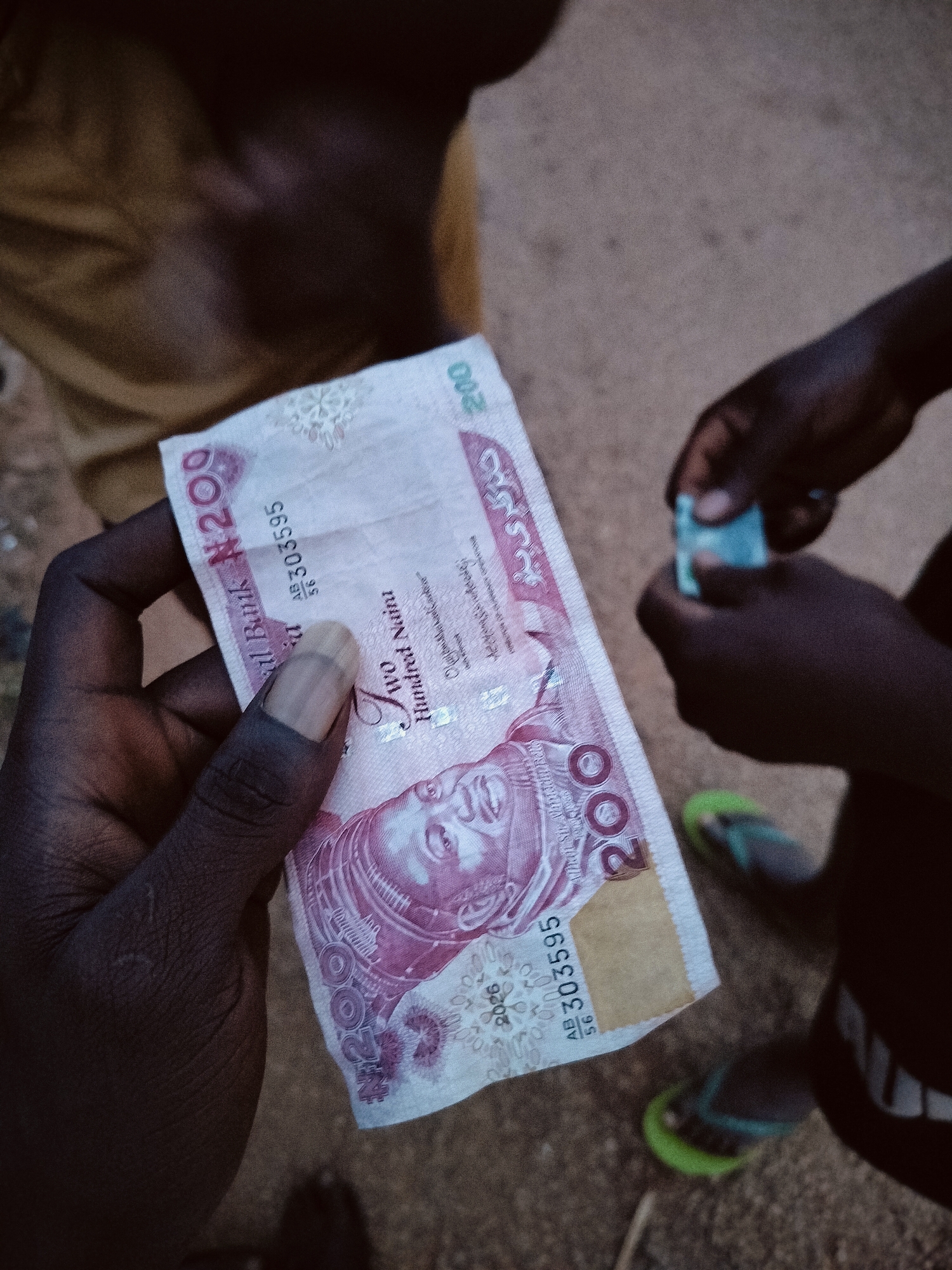 The image shows a close-up of a Nigerian 200 Naira banknote held in a person's hand. The banknote is pink and white and features a portrait of a woman. The serial number "AB 303595" is visible. In the background, another person's hands are visible, holding a small, blue object. The ground is sandy and appears to be outdoors. The lighting is dim, suggesting it might be evening or a shaded area. The image does not provide any specific information to identify its location within the city of Du, Nigeria.