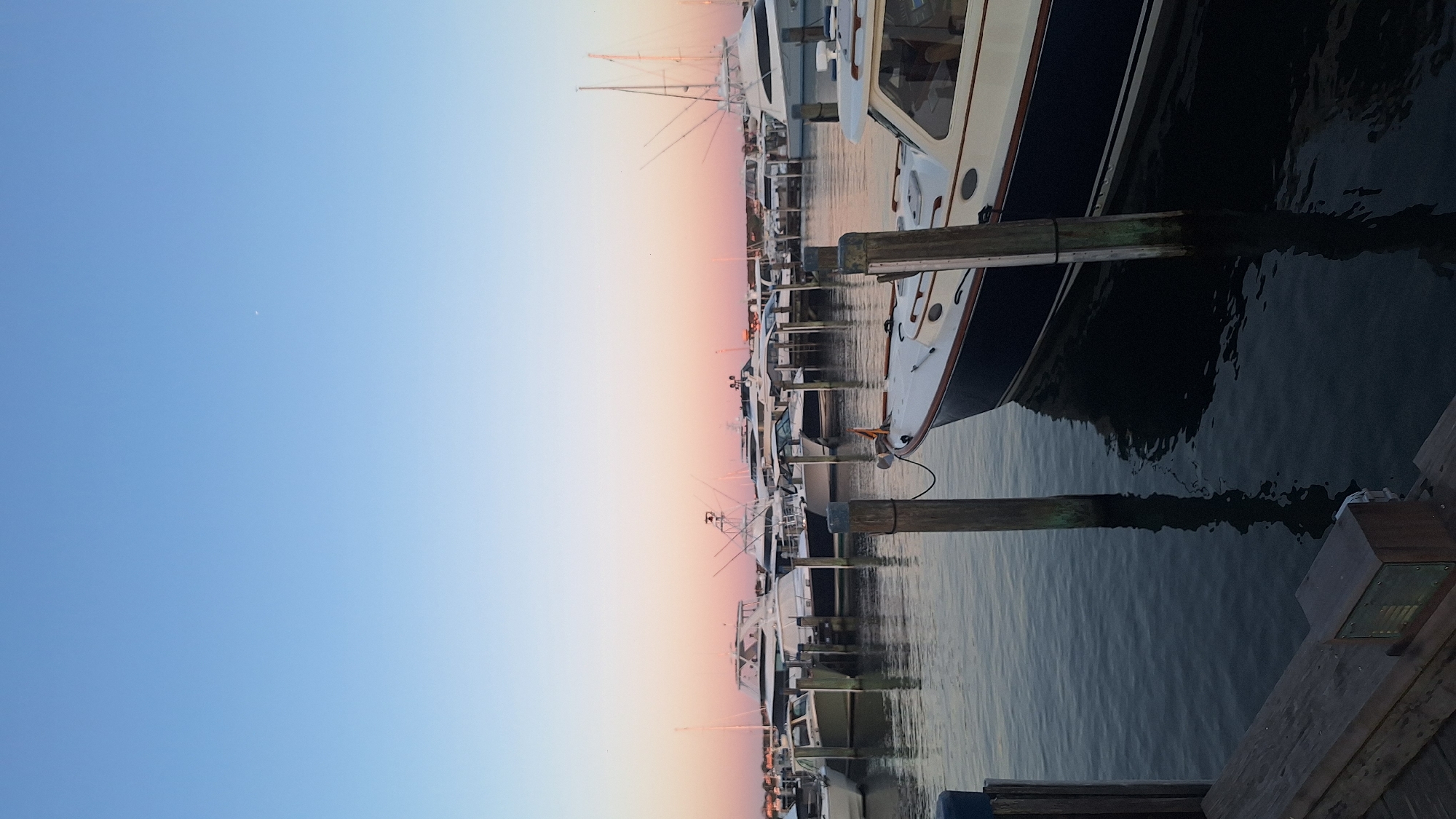 An outdoor, eye-level shot captures a marina scene in Nantucket, Massachusetts, USA. The water is calm and dark, reflecting the sky and boats. Several boats, ranging in size from small to large, are docked side-by-side. The boats are various colors, mostly white with some dark accents. The sky is a gradient of blue at the top, transitioning to a soft pink and orange hue near the horizon, suggesting it is either sunrise or sunset. A few faint clouds are visible in the upper portion of the sky. The marina appears quiet and serene, with no people visible. In the foreground, wooden pilings stand in the water, and part of a wooden dock is visible on the left side of the frame.