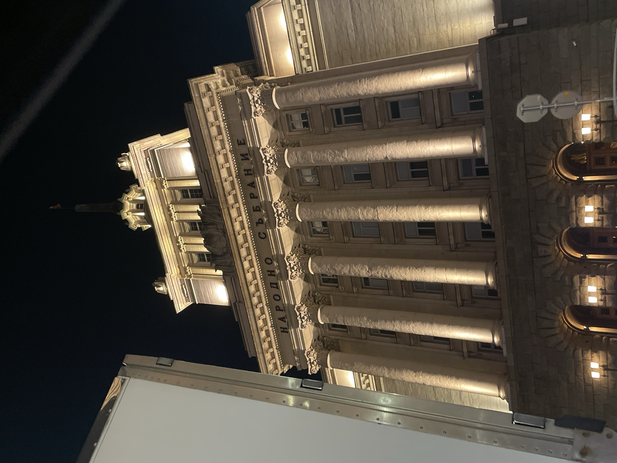The image shows the illuminated National Assembly of Bulgaria (Народно събрание) in Sofia, Bulgaria, at night. The building is neo-Baroque in style, and its facade features rows of columns. Part of what appears to be a white truck or vehicle is visible in the foreground at the bottom of the picture. The sky is dark.