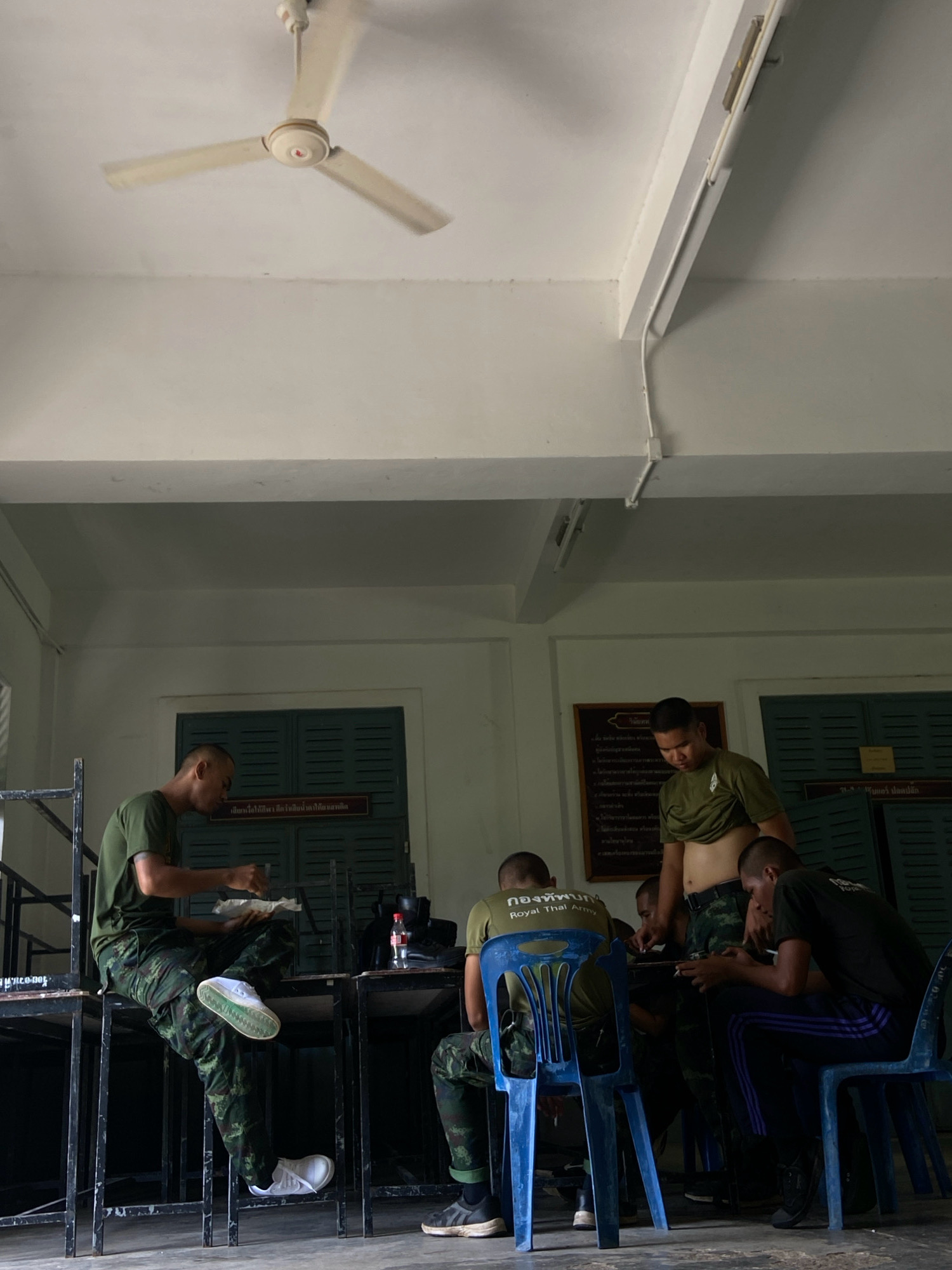 The image depicts a group of soldiers in what appears to be a common area, possibly a barracks or mess hall, within Ban Si Yaek San Nare San, Thailand. The room has a concrete floor and white walls, with dark green louvered shutters on the windows. A white ceiling fan is visible overhead, suggesting a warm climate.
Several soldiers, dressed in camouflage uniforms, are seated and interacting. One soldier on the left is seated on a metal structure, holding a white container and what seems to be food, and is looking down. Another soldier is sitting in a blue plastic chair, looking at a phone. A third soldier, standing and wearing a green t-shirt with his midriff exposed, is looking down at a table where other soldiers are gathered. Two other soldiers are seated at a table, one of whom is actively using a phone.
The overall atmosphere suggests a break or downtime for the soldiers. The presence of individual phones indicates some level of personal access and leisure. The environment appears functional and utilitarian, consistent with military facilities. A framed sign with Thai text is mounted on the wall behind the standing soldier, but its content is not discernible.