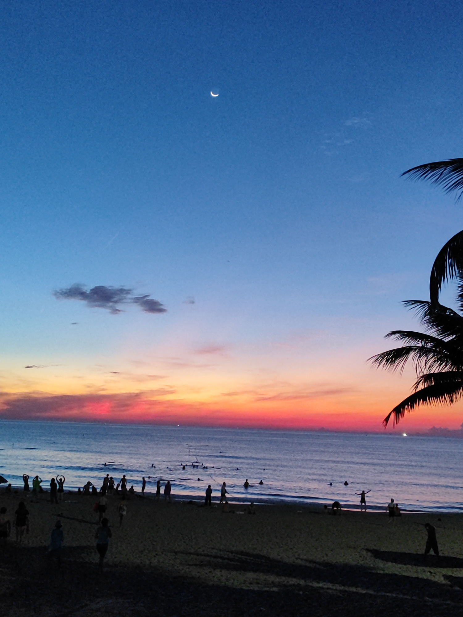 A dusk scene at My Khe Beach in Đà Nẵng, Vietnam, captures the transition from day to night. The sky, a gradient of deep blue at the top to vibrant orange and pink near the horizon, is adorned with a crescent moon and wispy clouds. The ocean is calm, with gentle waves lapping the shore. On the sandy beach, a diverse group of people engage in various activities. Some are standing and interacting, while others are seated or walking along the shoreline. A few individuals are visible in the water, suggesting swimming or wading. Silhouettes of palm trees frame the right side of the image, adding a tropical touch to the landscape. The overall atmosphere is one of relaxed enjoyment and the serene beauty of a coastal sunset.