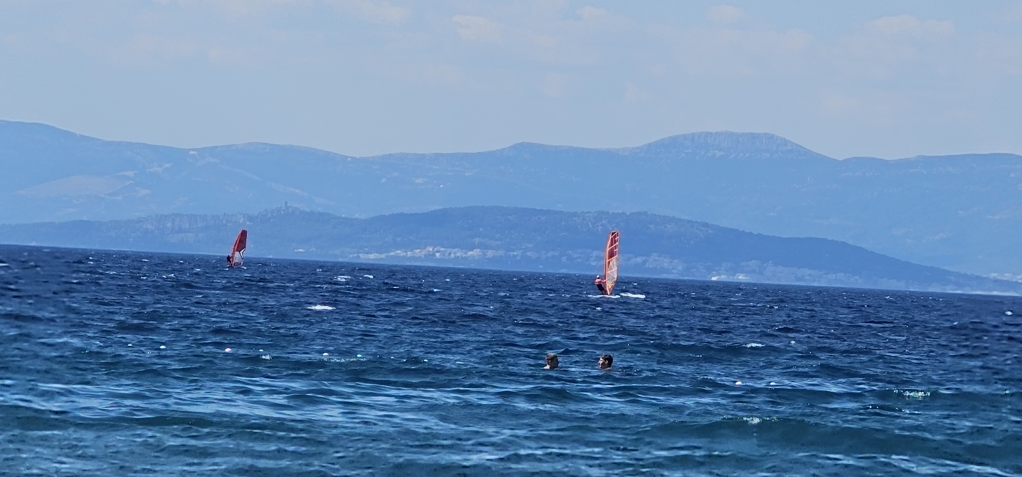 In the city of Supetar, Croatia, on a clear day with a sky of pale blue and scattered clouds, the Adriatic Sea is choppy and a deep blue. Two windsurfers are visible on the water, one with a red and white sail closer to the left side of the frame, and another with a red and orange sail on the right. Between them, closer to the foreground, three people are swimming with their heads visible above the water. In the background, layers of blue-toned mountains rise up, with the faint outlines of a town or village visible on the slopes of one of the closer mountain ranges. The overall scene depicts a recreational day at sea.