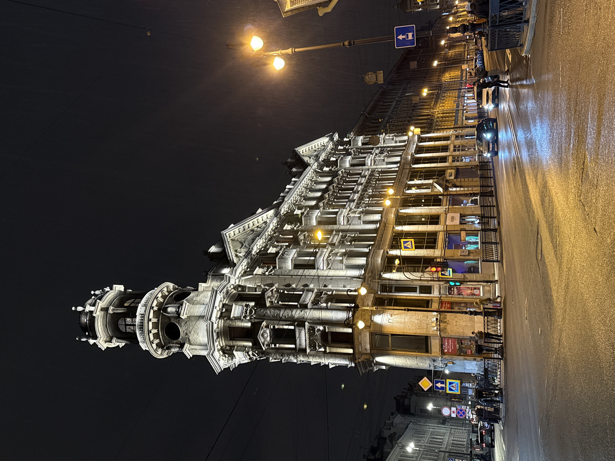 The image depicts a nighttime street scene in Saint Petersburg, Russia. An ornate, multi-story building is prominently featured, illuminated with bright lights. The building has a tower structure on one end. The street is wet and reflects the light from street lamps and building lights. People are walking along the sidewalk, and cars are driving on the street. Street signs and traffic signals are visible. The sky is dark and clear.