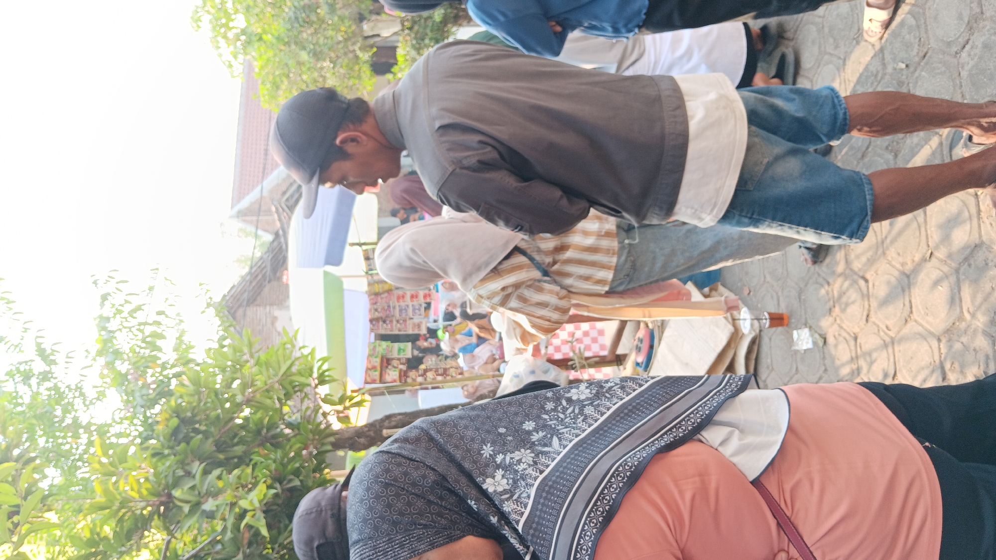 In the city of Cerme, Indonesia, a group of people are gathered outdoors, possibly at a market or street fair. In the foreground, a person is partially visible from the side, wearing a peach-colored shirt and a patterned hijab. To their right, a man in a dark jacket and denim shorts stands with his arms crossed, looking to the side. Further back, other individuals, some wearing hijabs and traditional Indonesian attire, are browsing goods displayed on stalls. The environment appears to be a sunny day, with sunlight filtering through trees and casting shadows on the paved ground. The background shows simple buildings and tents, suggesting a local community gathering.